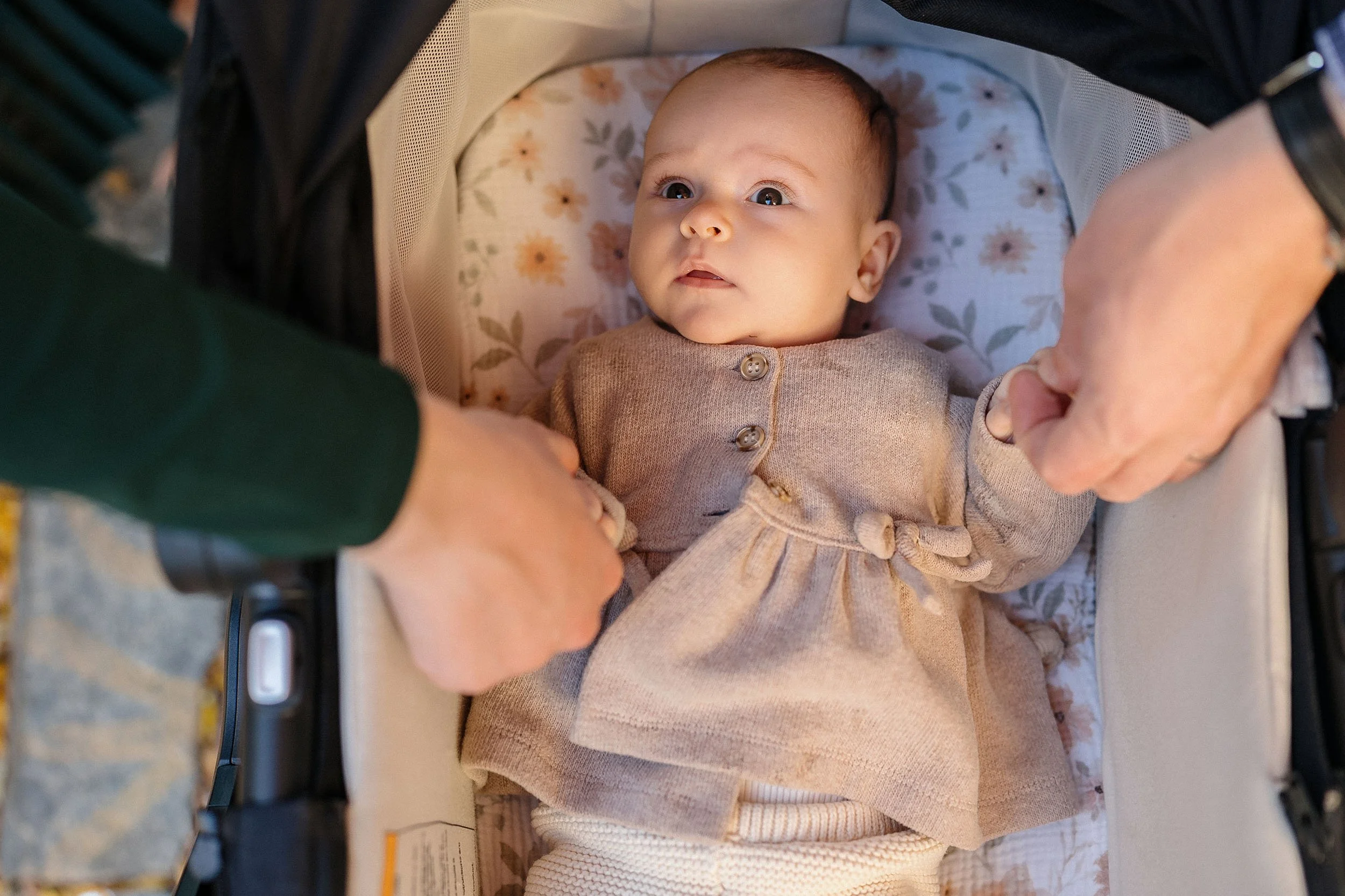 A baby girl lying in a beige car seat, looking upward with a curious expression, being held gently by two adult hands.