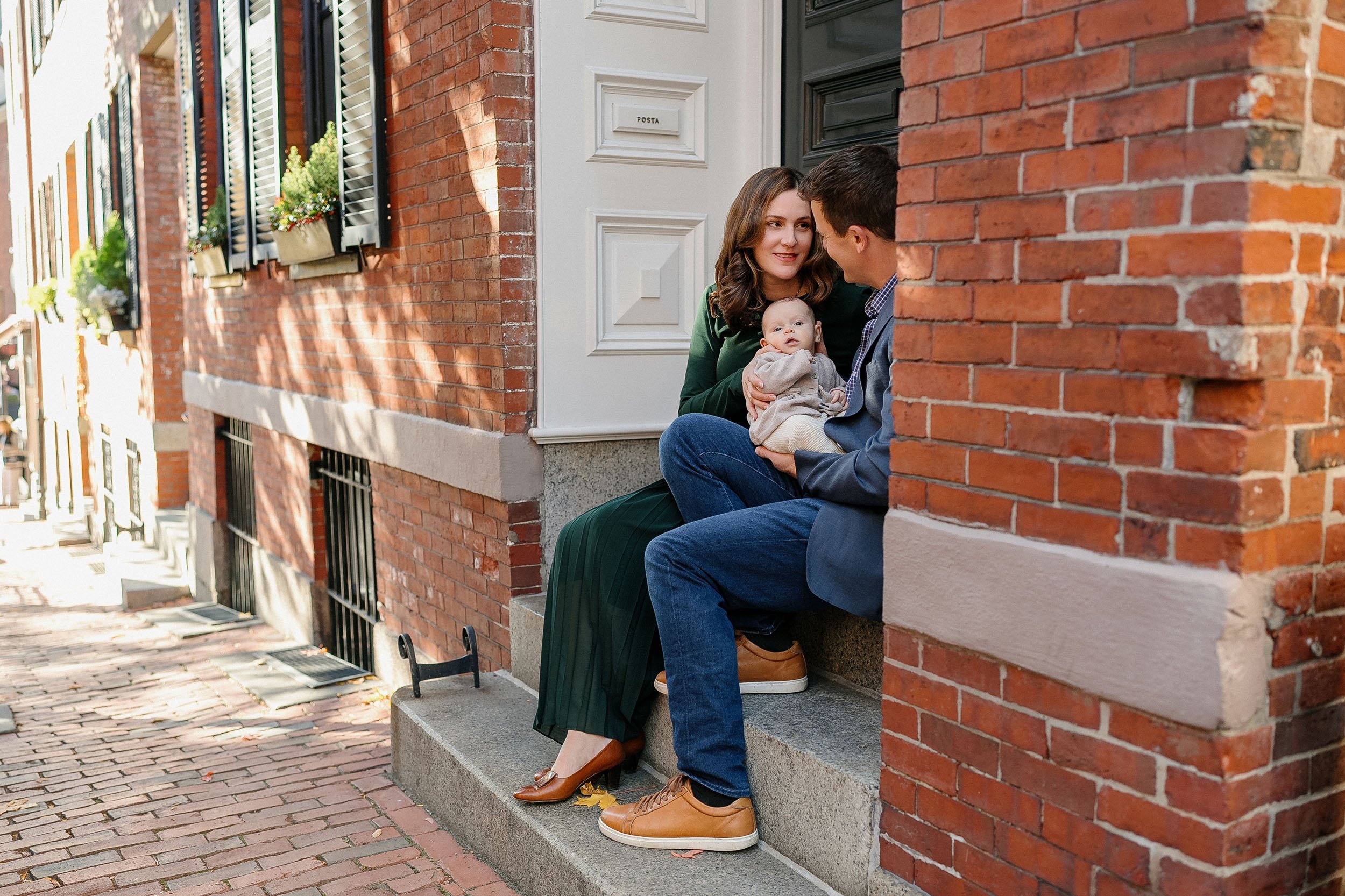 A couple and a baby sitting on a front porch steps of a brick house talking and smiling at each other.