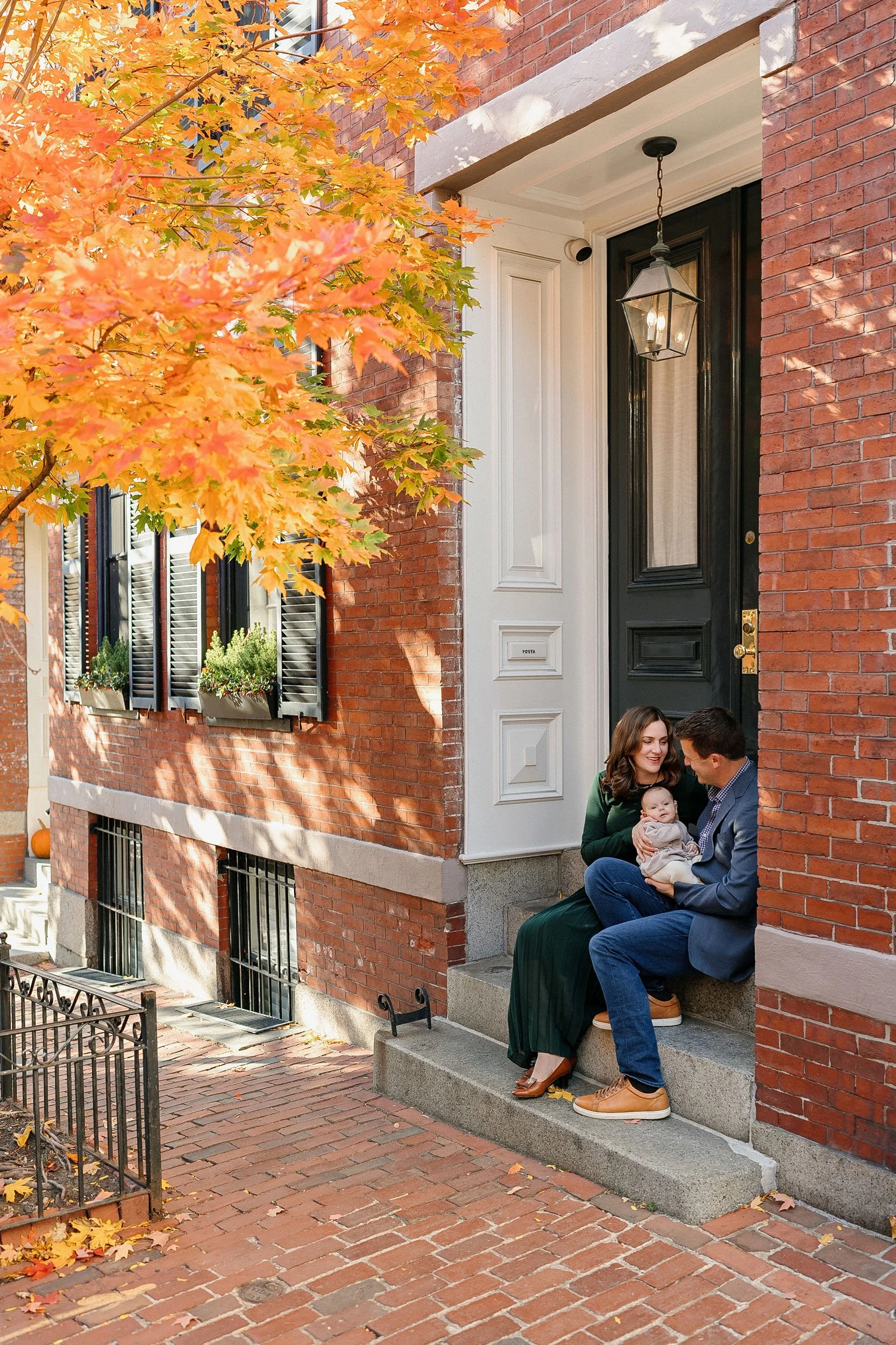 A happy couple sitting on the front steps of a brick house, holding their baby. The woman is wearing a dark green dress and the man is dressed in a blue blazer and jeans. There is an orange and yellow autumn tree overhead, and window boxes with plant