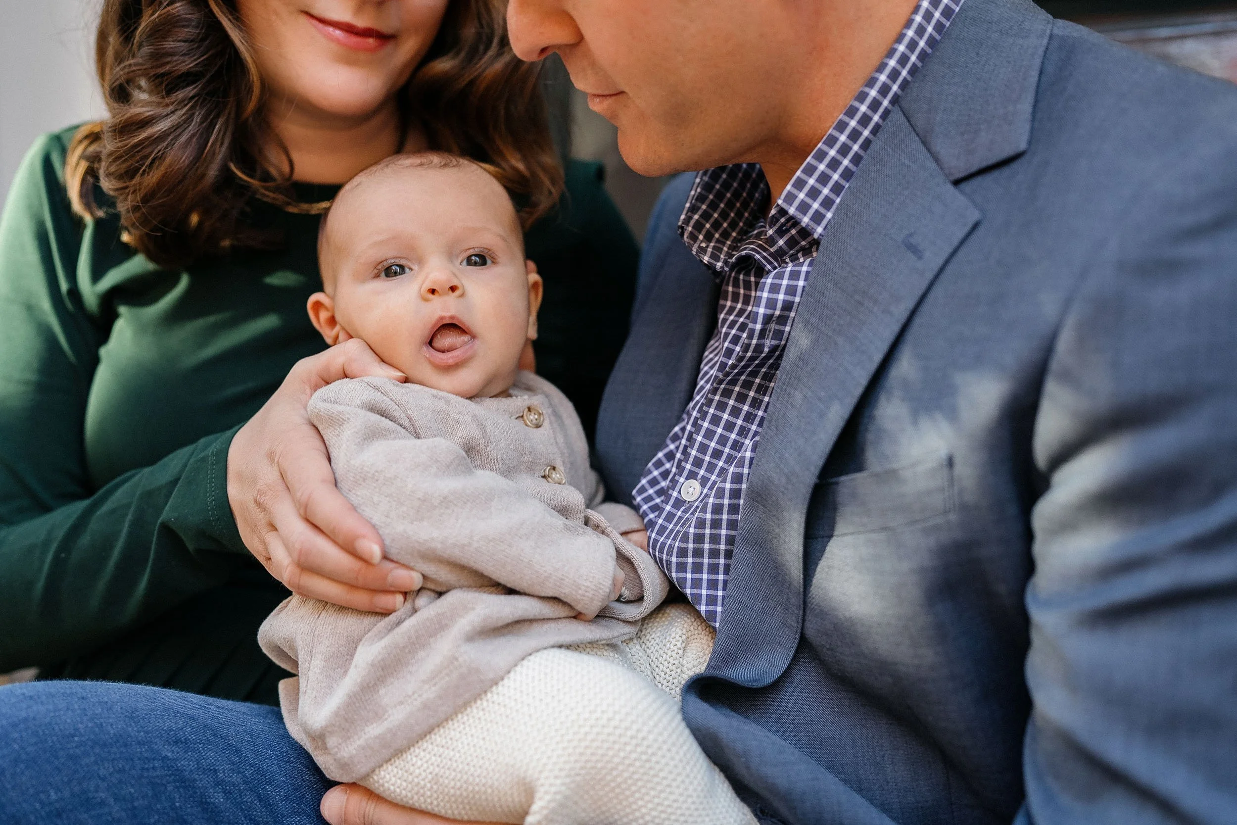 A family with a woman, a man, and a baby, sitting close together. The baby has wide eyes and an open mouth, and is being gently held by the woman.