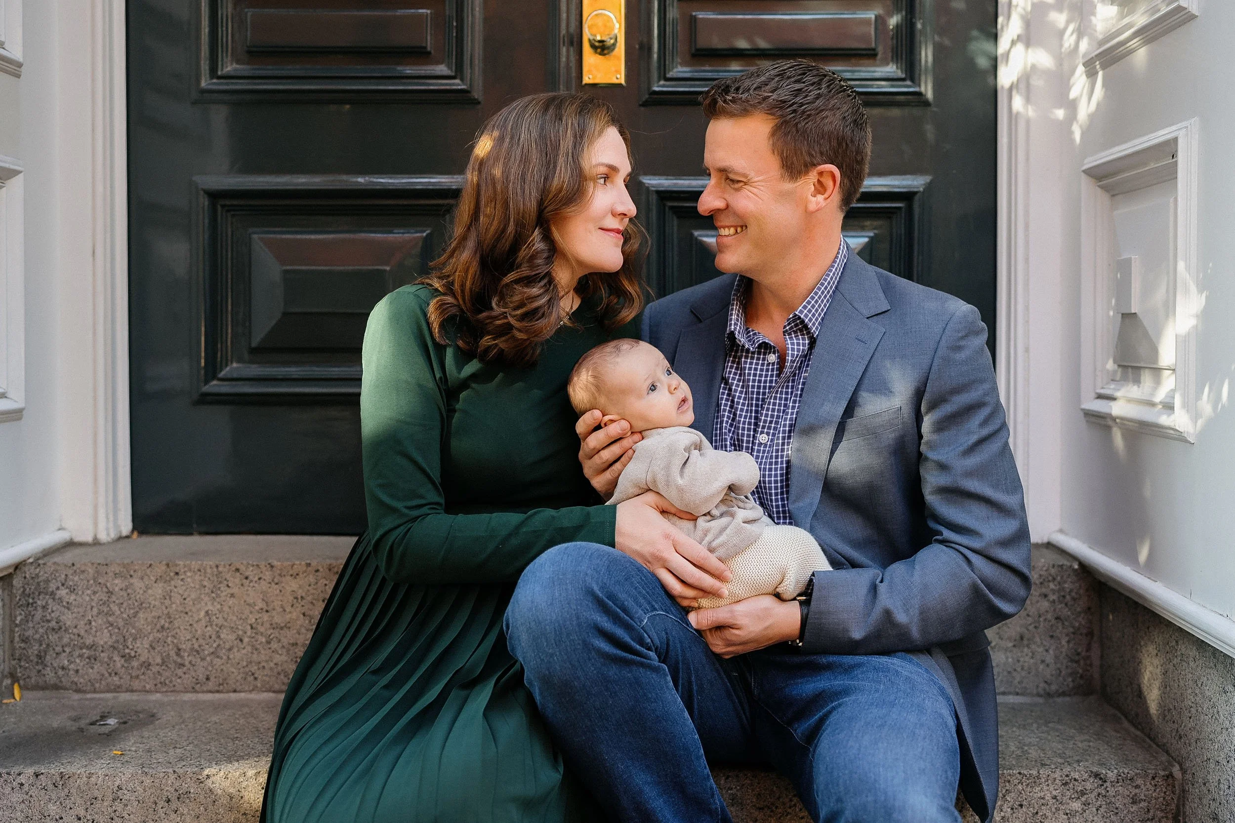 A smiling family of three sitting on front porch steps. The mother and father are looking at each other, holding their baby, who is gazing upward. The mother has brown hair and is wearing a green dress. The father has short brown hair and is wearing 