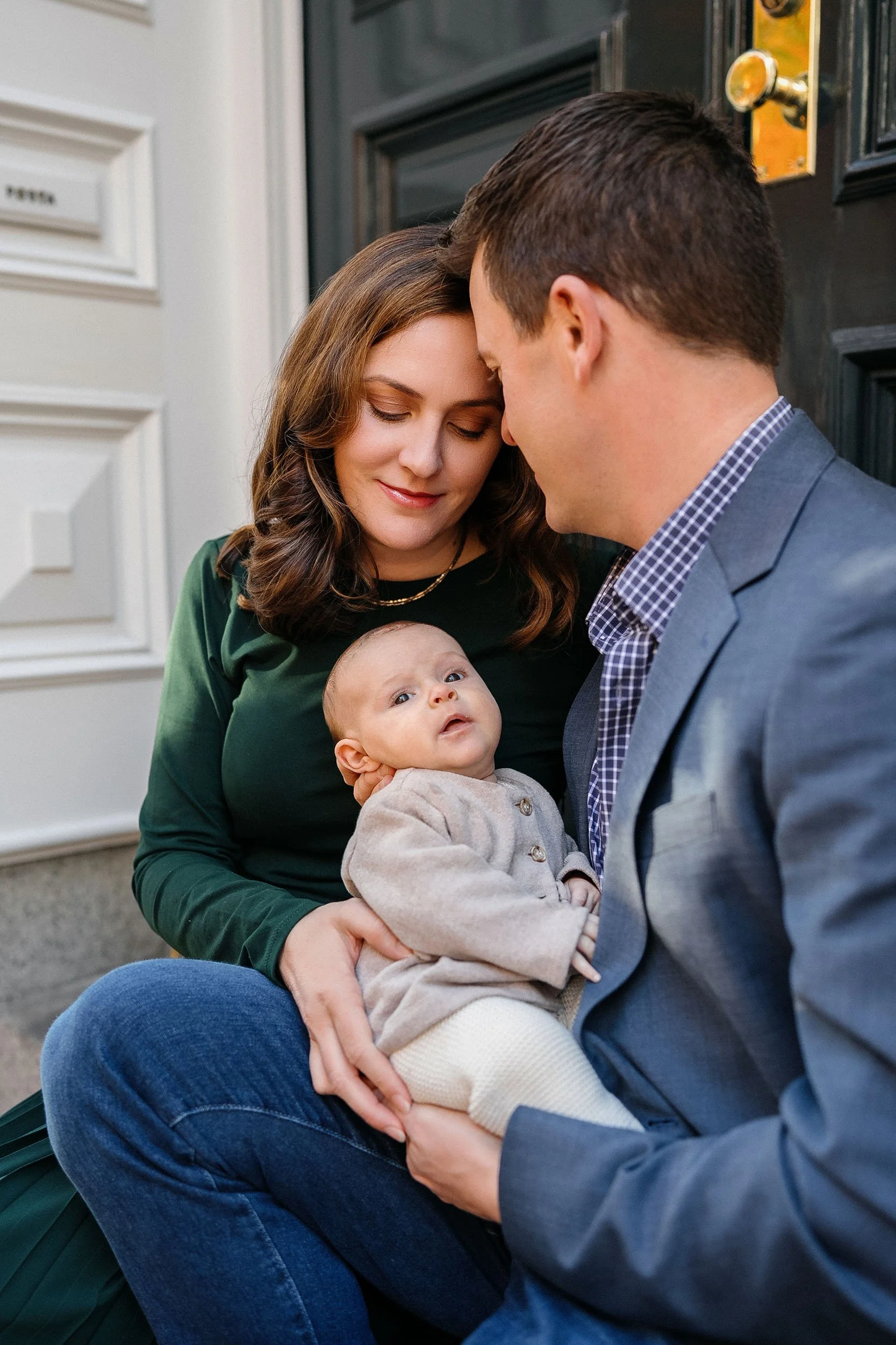 A family of three sits on front steps in front of a black door, with the mother holding the baby and the father leaning in close, all peacefully smiling.