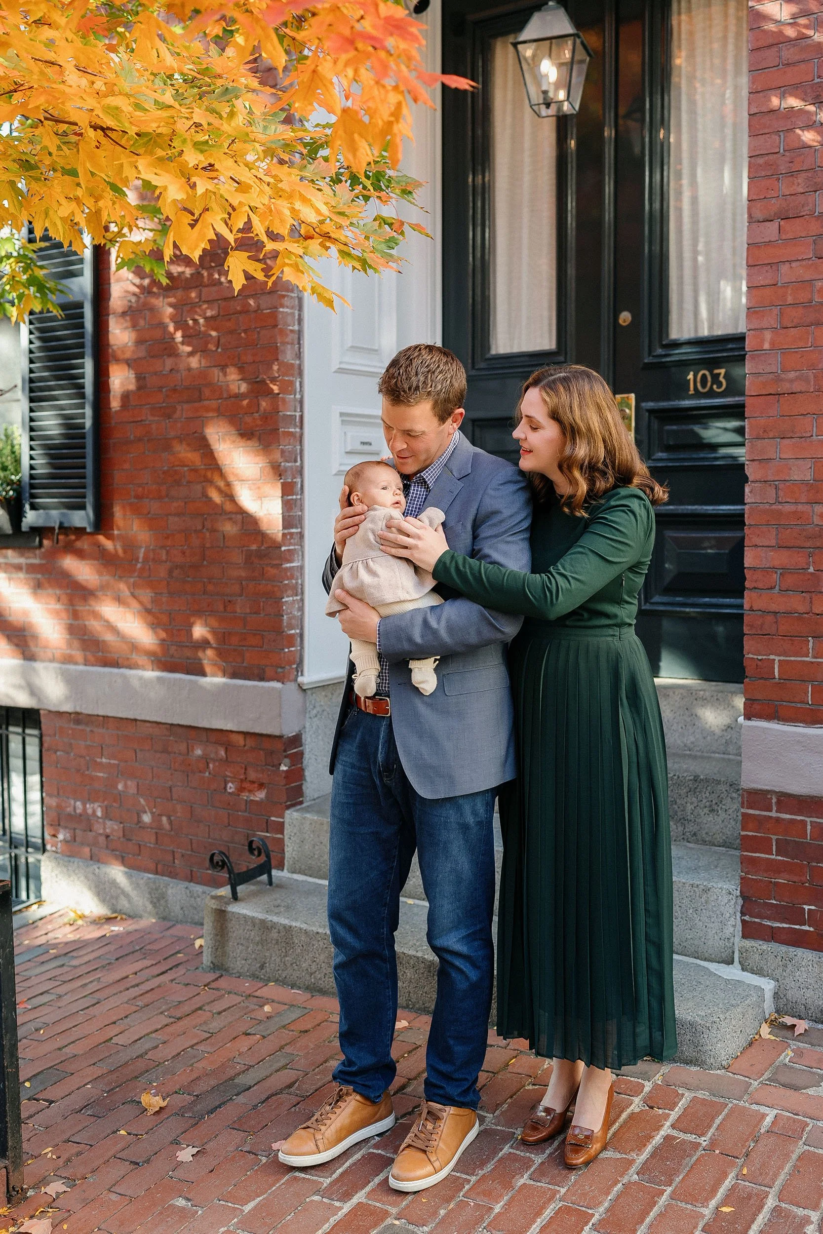 A family of three, a man, woman, and baby, standing on a brick sidewalk in front of a black door with house number 103, and a brick house with a window with open shutters, during fall with orange and yellow leaves on a tree.