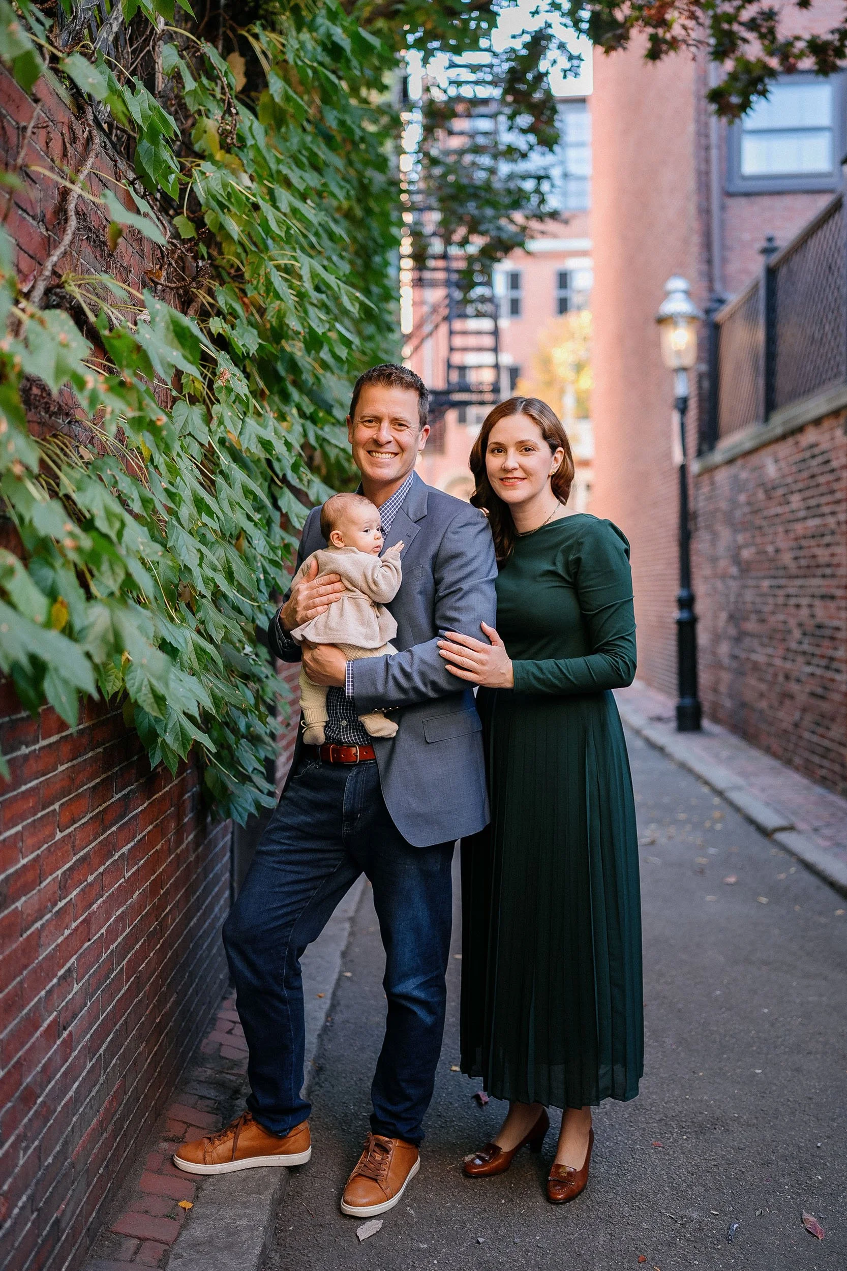 Family of three, father, mother, and baby, standing on alleyway with brick walls and ivy, smiling for the camera.