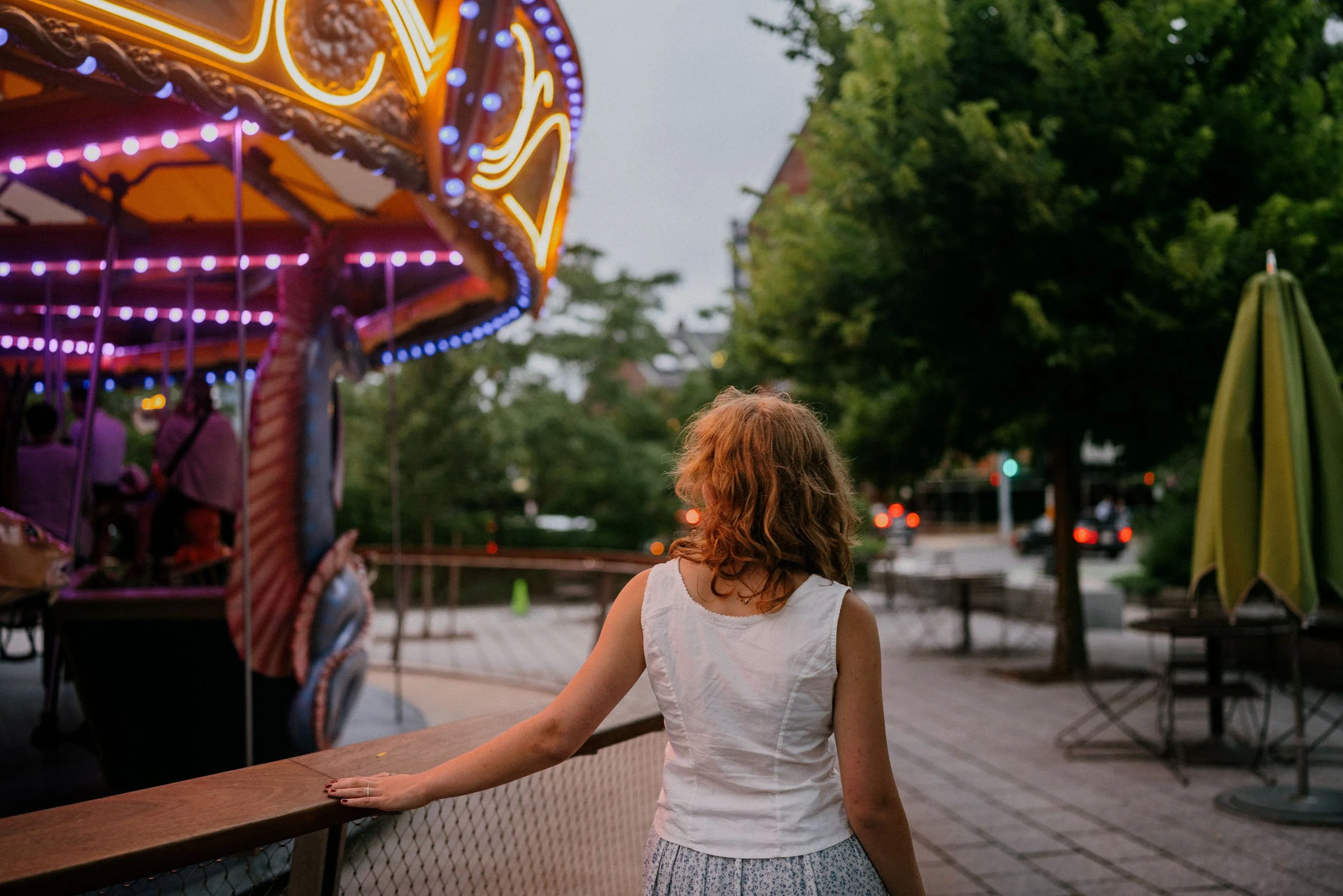 A woman with curly hair wearing a white sleeveless top standing near a wooden fence at an amusement park with a lit-up carousel in the background during dusk.