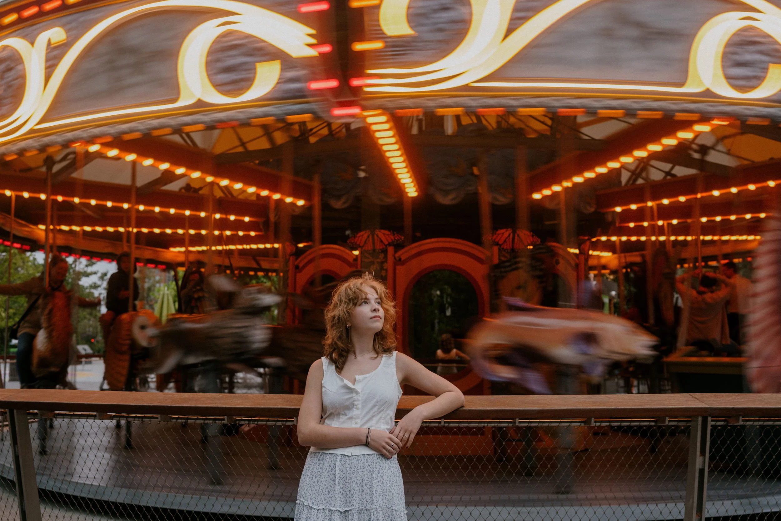 A young girl with curly red hair wearing a white dress standing in front of a carousel at an amusement park, with the carousel in motion and blurry lights.