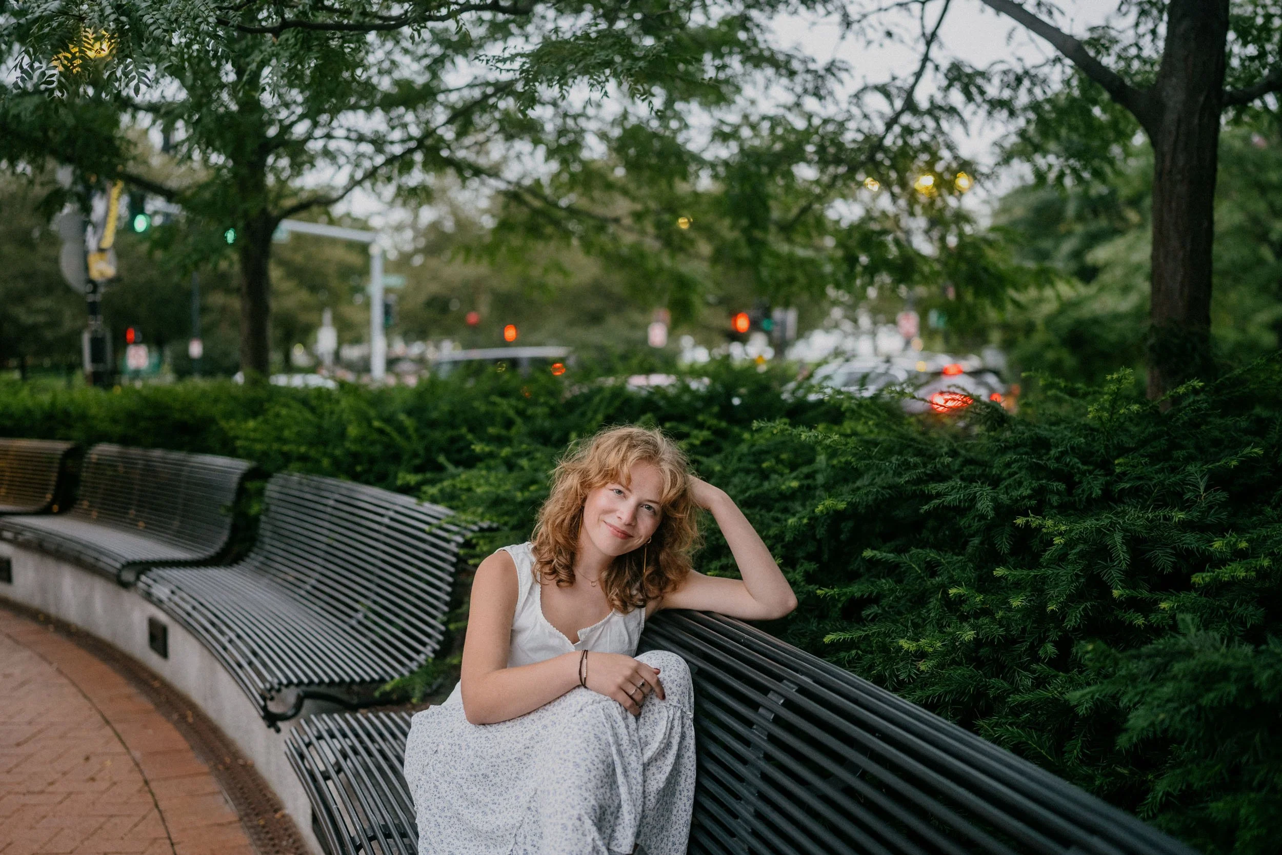 A young woman with curly red hair sitting on a park bench surrounded by green foliage, during the evening.