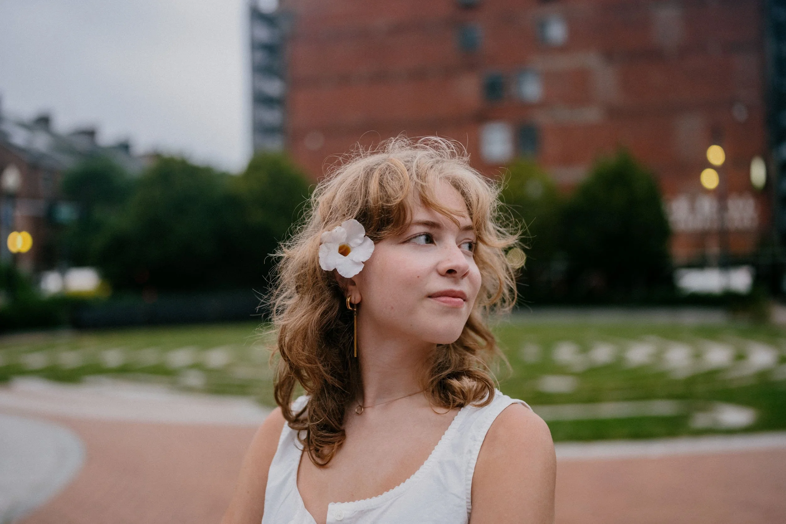 A young woman with curly blonde hair and a white flower in her hair, wearing a white sleeveless top, standing outdoors in a park with a brick building and trees in the background.