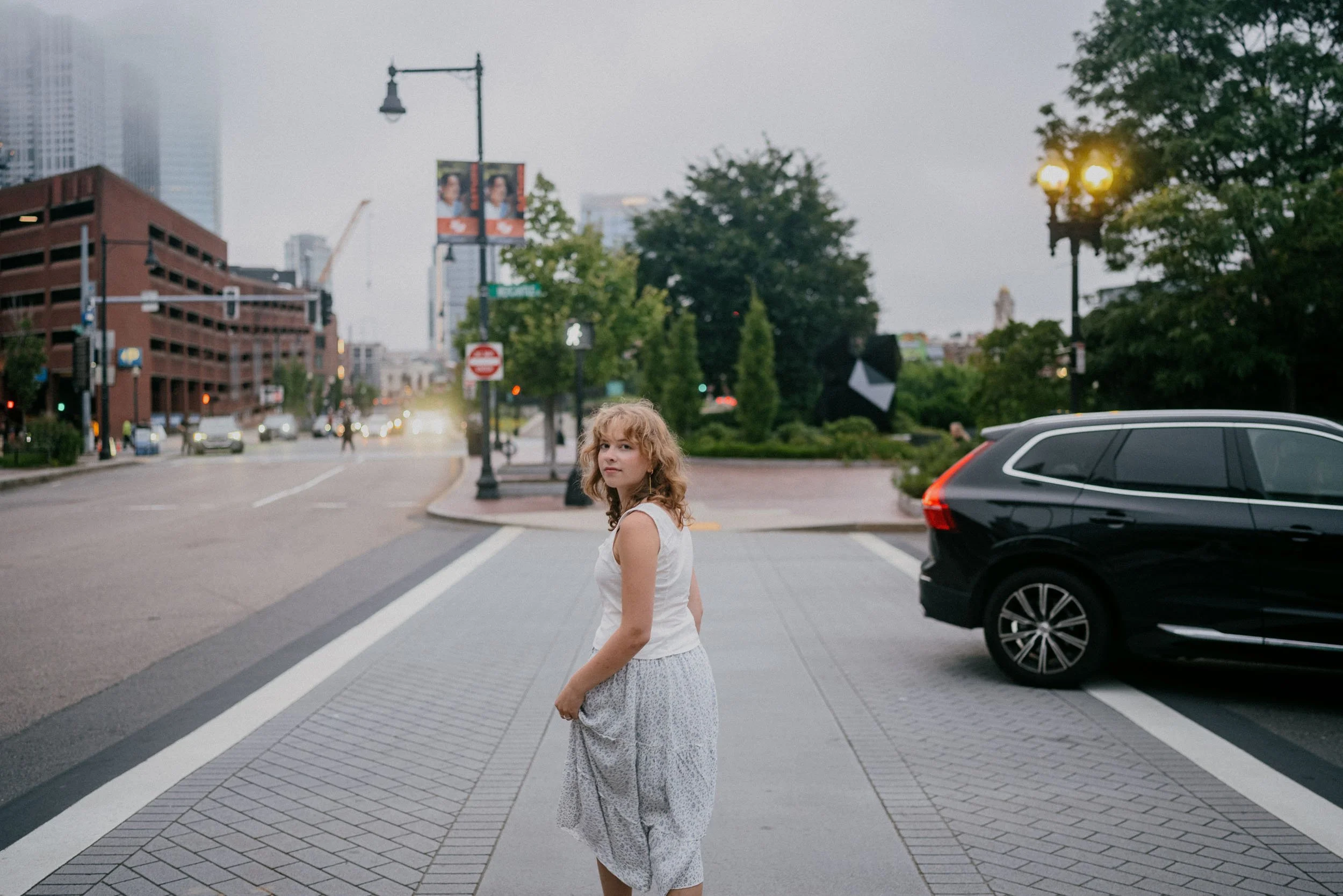 A young woman with curly blonde hair wearing a white sleeveless top and a long patterned skirt standing on a city sidewalk at dusk, with buildings, trees, a black car, and streetlights in the background.