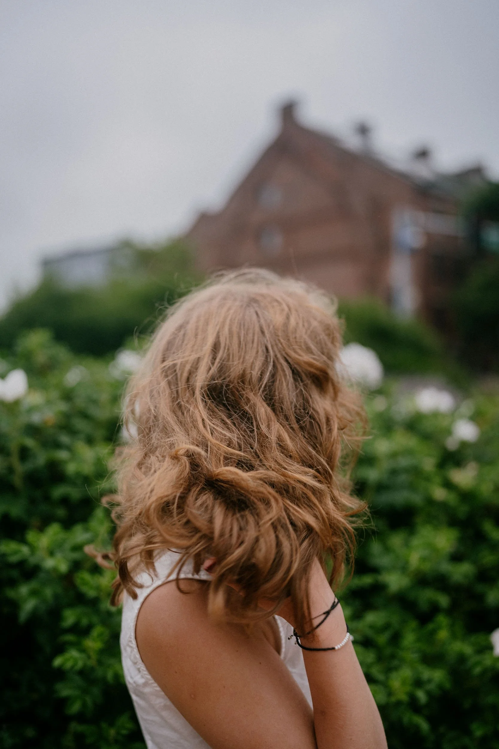 A woman with curly red hair standing outdoors among greenery and white flowers, facing away from the camera with a house in the blurred background.