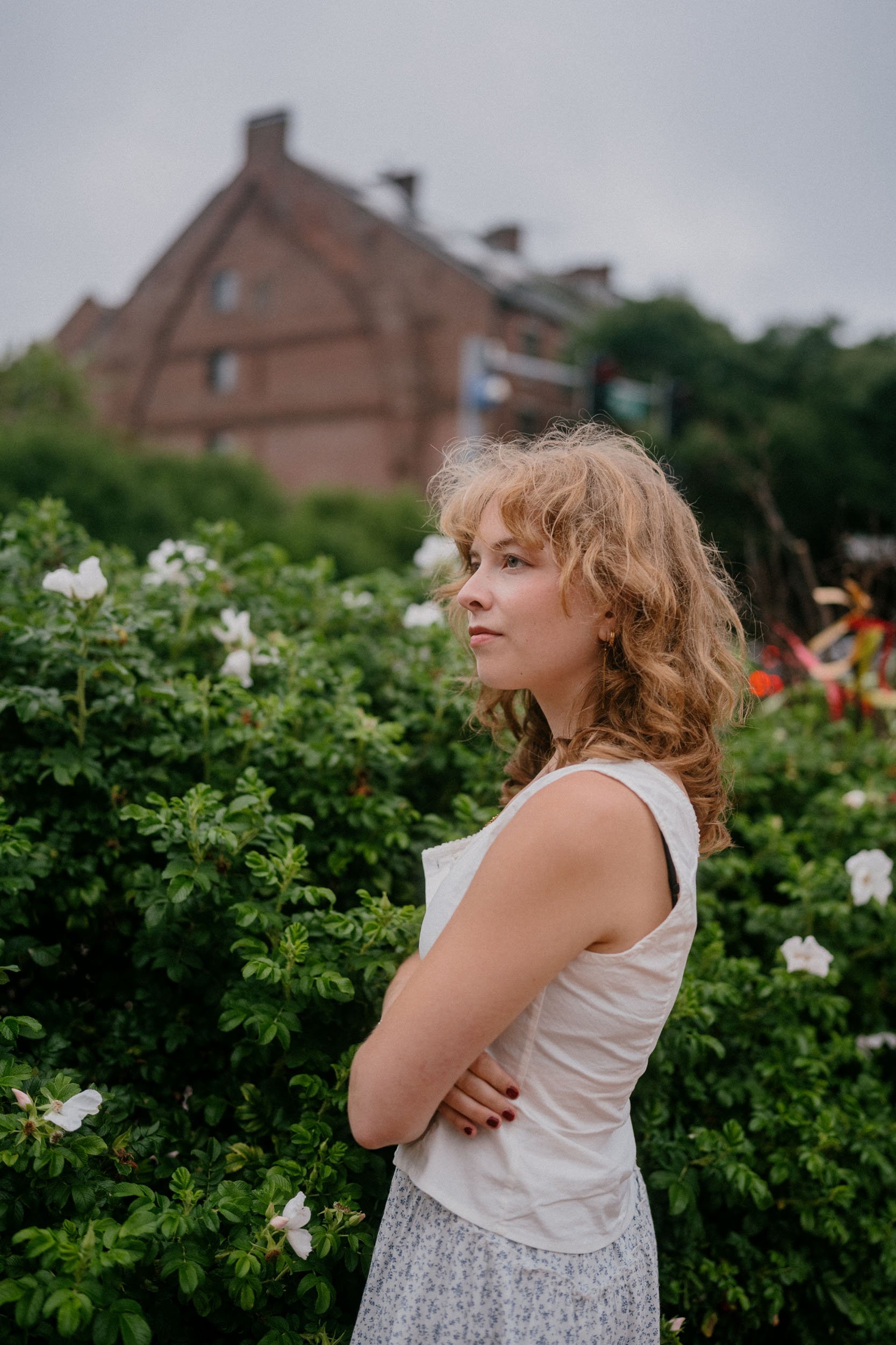 A woman with curly blonde hair standing with her arms crossed among green bushes with white flowers, facing right, with a large brick house in the background.