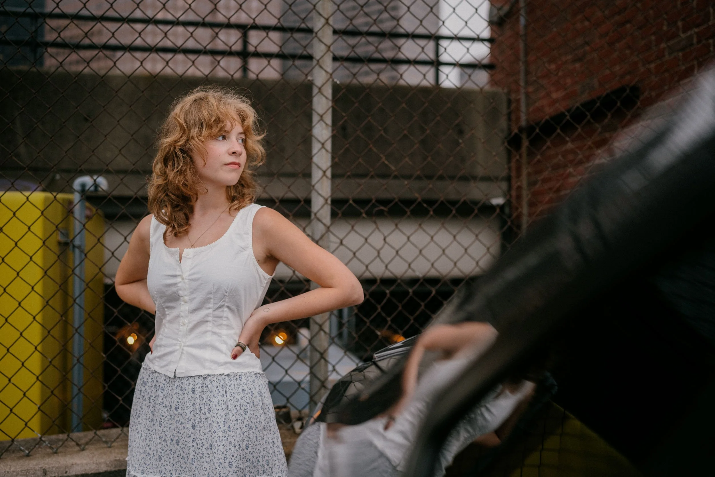 A woman with curly red hair wearing a white sleeveless top and a patterned skirt standing with her hands on her hips near a chain-link fence in an urban setting.