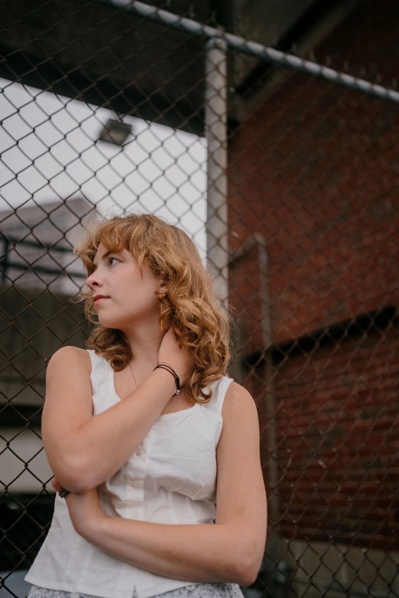 A young woman with curly red hair and light skin stands near a chain-link fence, looking to her left with her hand touching her neck, in an urban setting with brick and metal structures in the background.