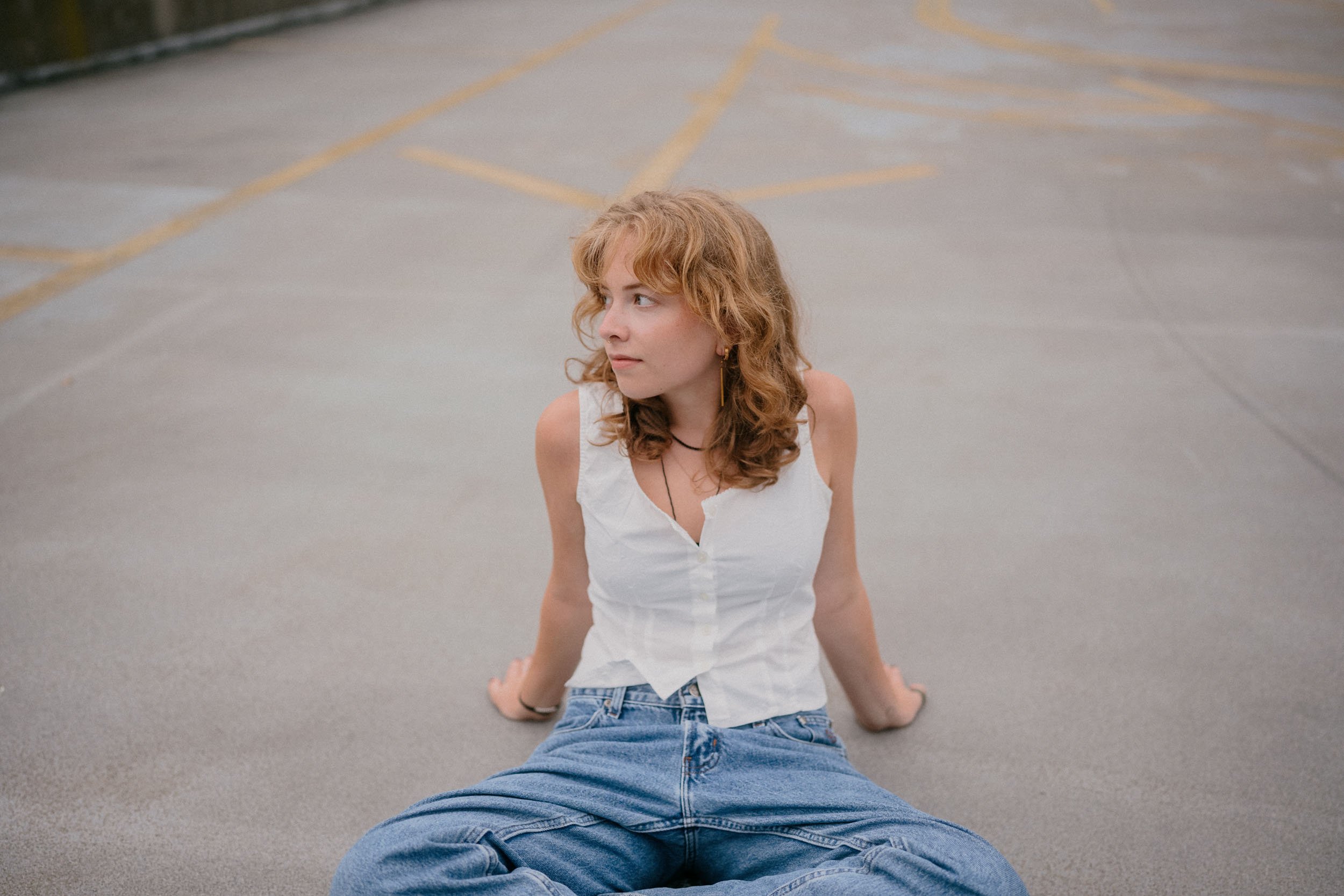Young woman with curly red hair sitting on a parking lot ground, looking to her left, wearing a white sleeveless top and blue jeans.