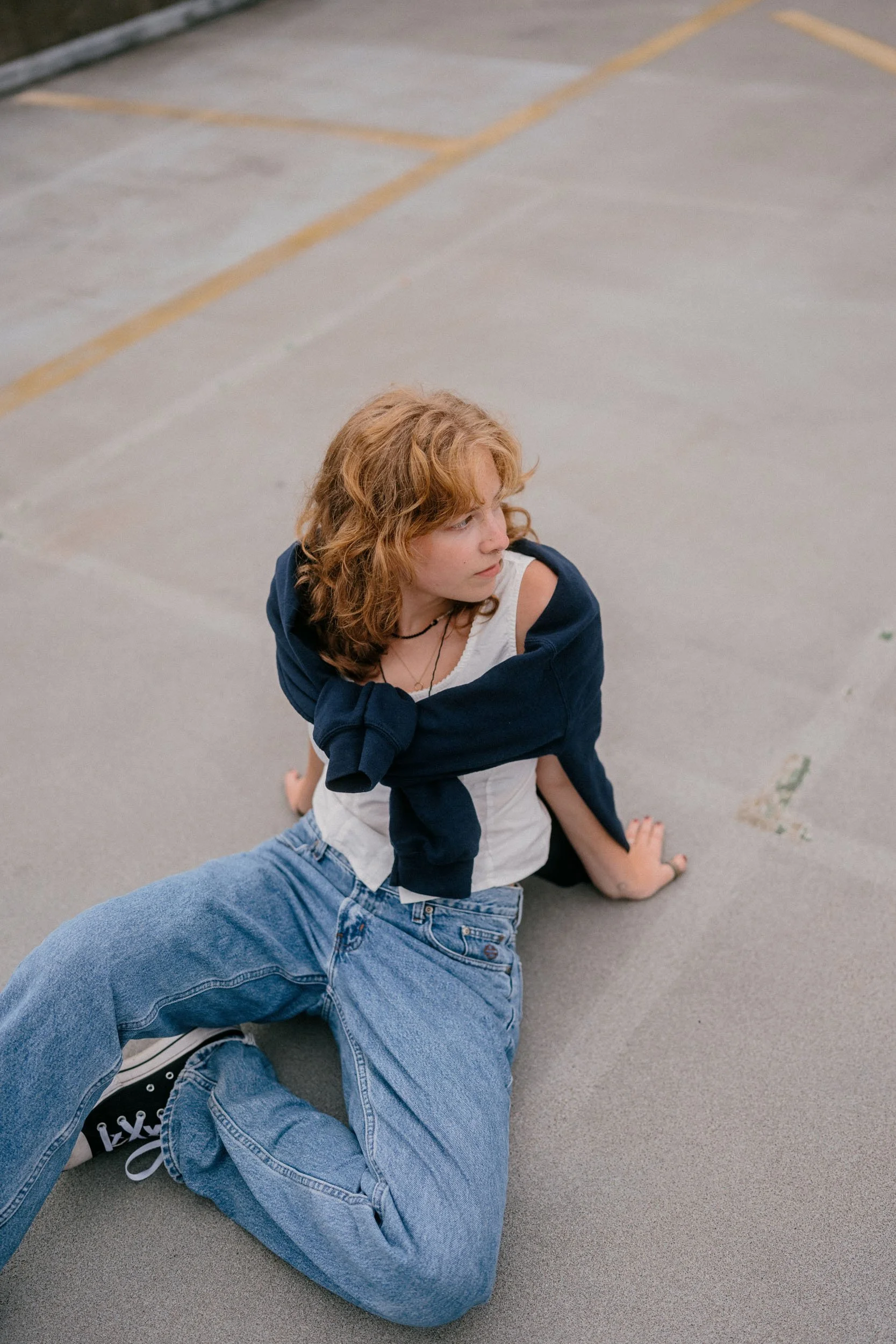 A young woman with curly red hair sitting on the ground in an empty parking lot, looking off to the side with a thoughtful expression, wearing a white top, blue jeans, black sneakers, and a navy blue sweatshirt draped over her shoulders.