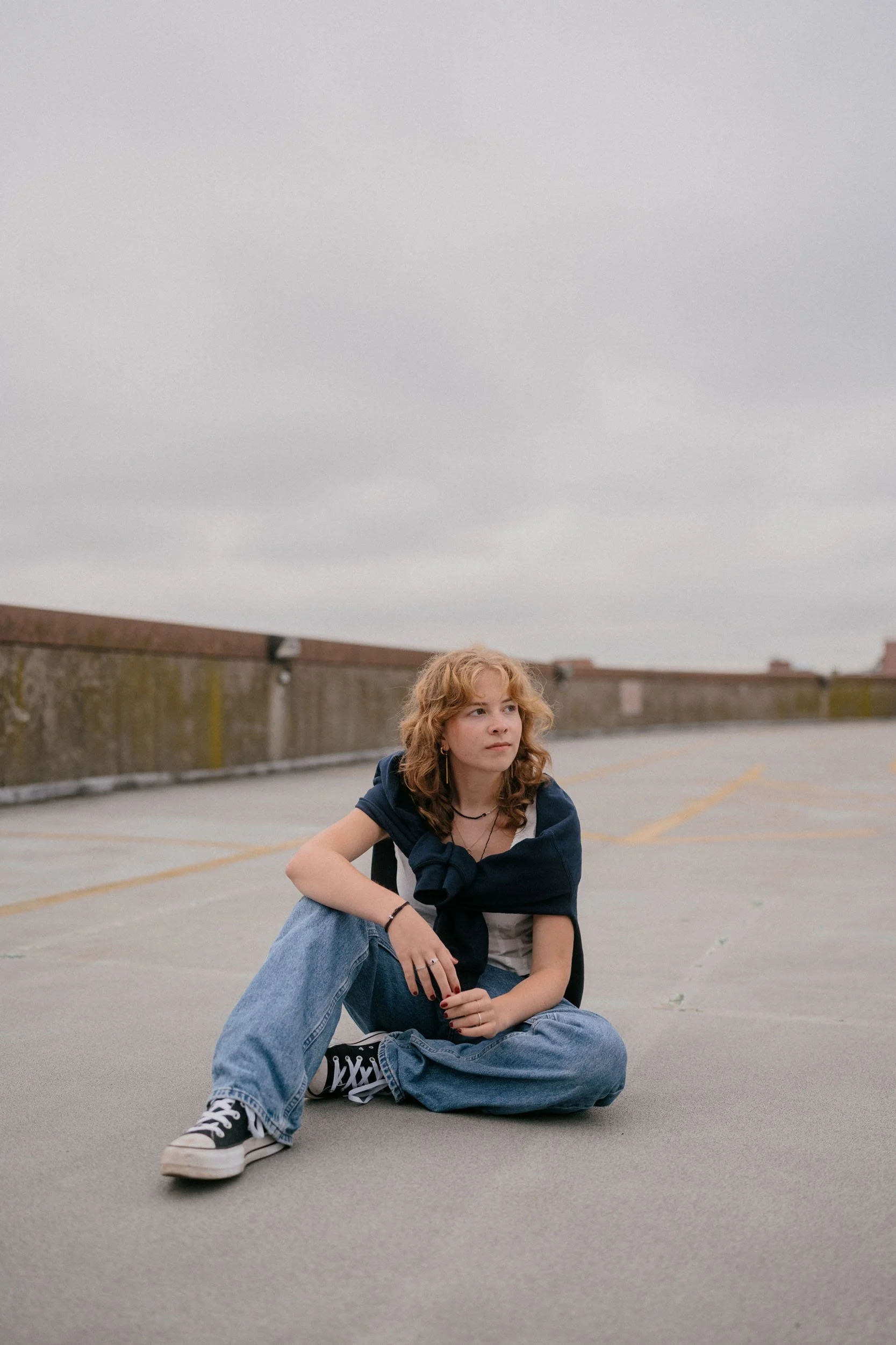 A young woman with curly red hair sitting on the ground in an empty parking lot, looking thoughtfully to the side, with a grey cloudy sky overhead.
