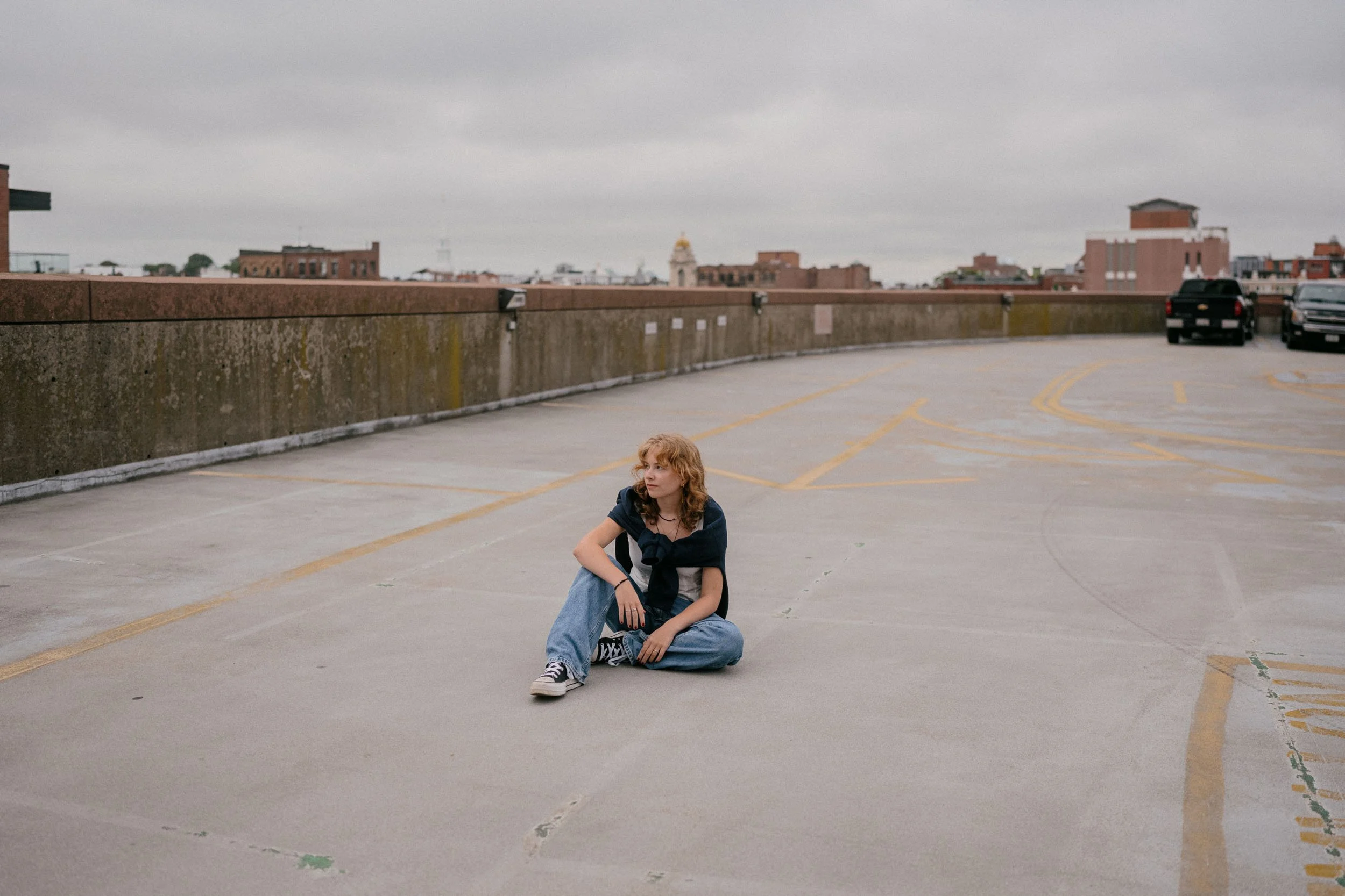 A young woman with curly red hair sitting on the ground in an empty rooftop parking lot on a cloudy day, with city buildings in the background.