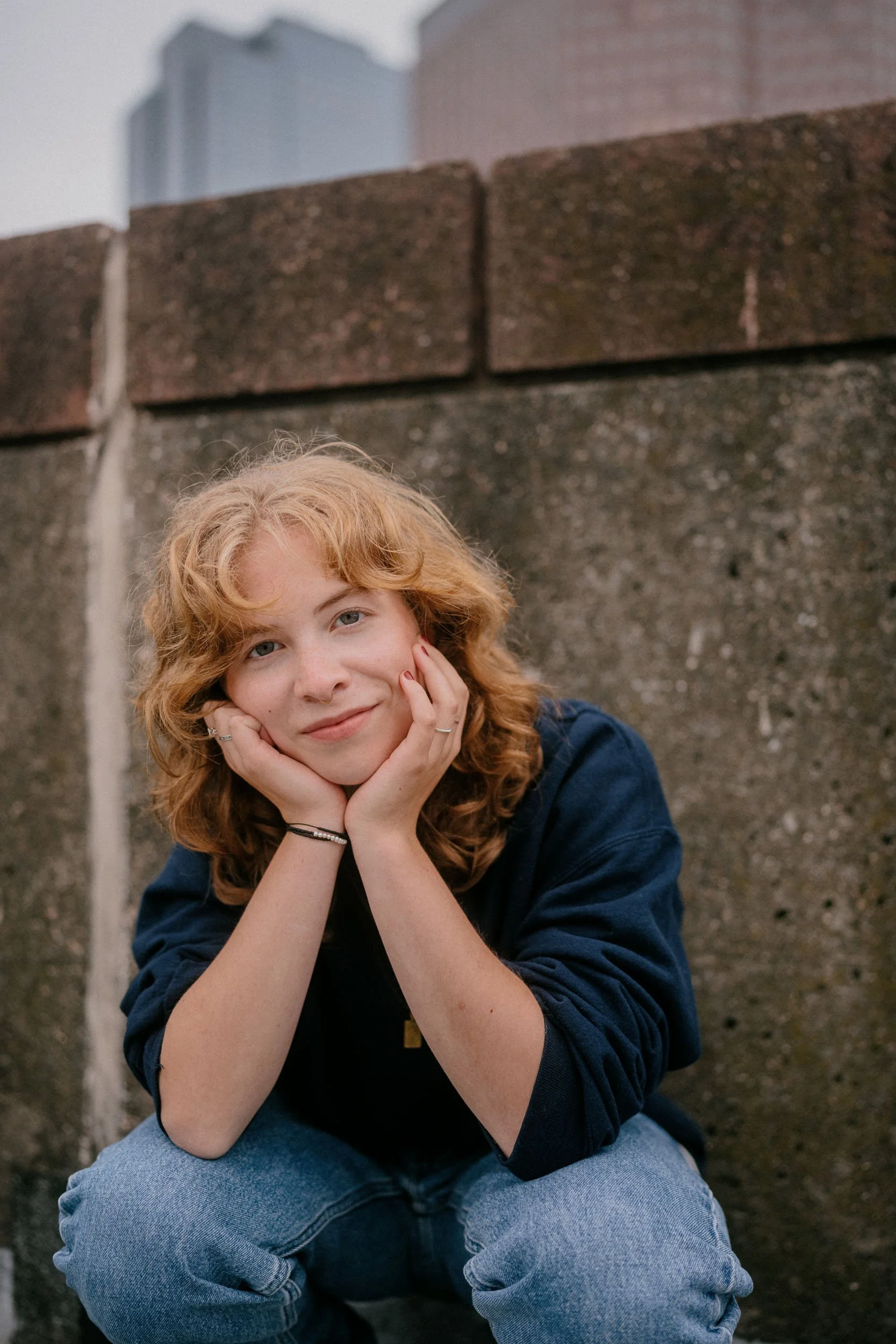 A young woman with curly red hair sitting outdoors against a brick wall, resting her chin on her hands, smiling gently at the camera.