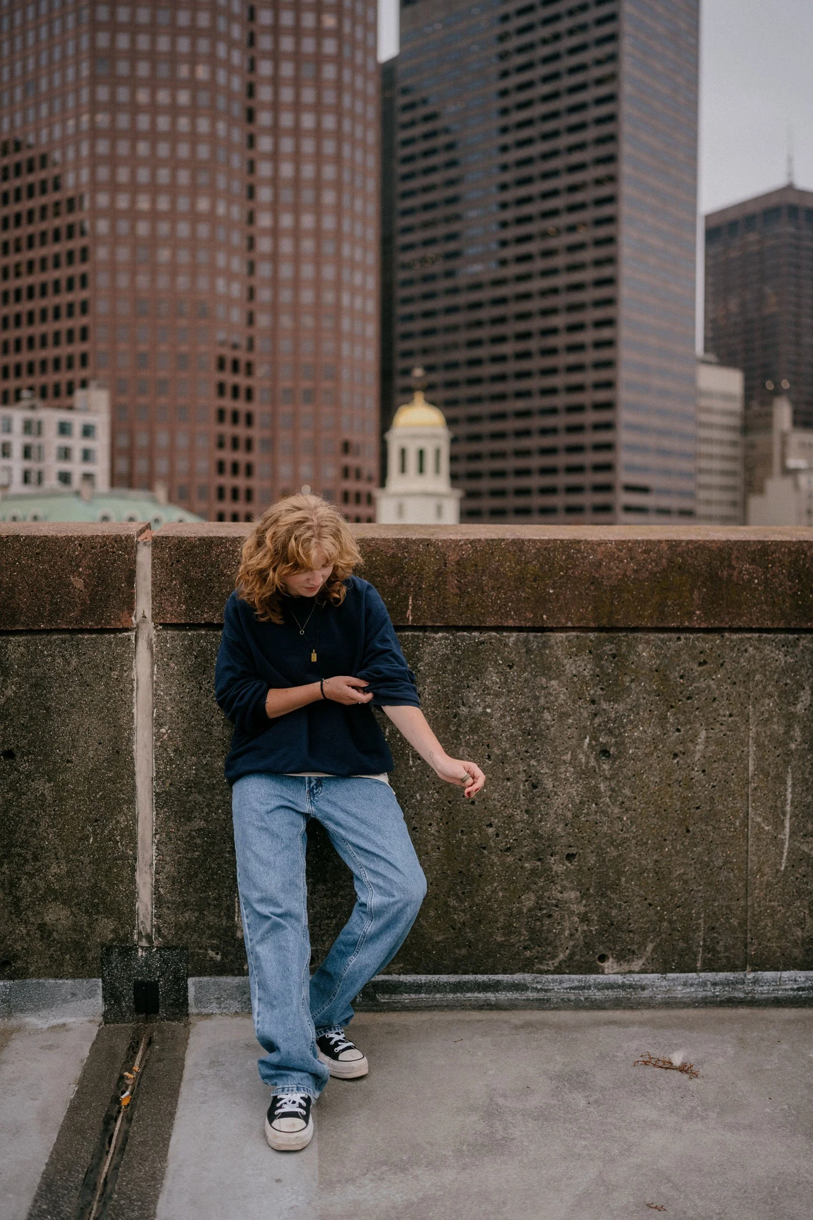 A person with curly blonde hair wearing a navy sweater, light blue jeans, and black sneakers stands against a concrete wall on a rooftop with a city skyline in the background, including tall skyscrapers and a building with a yellow dome.