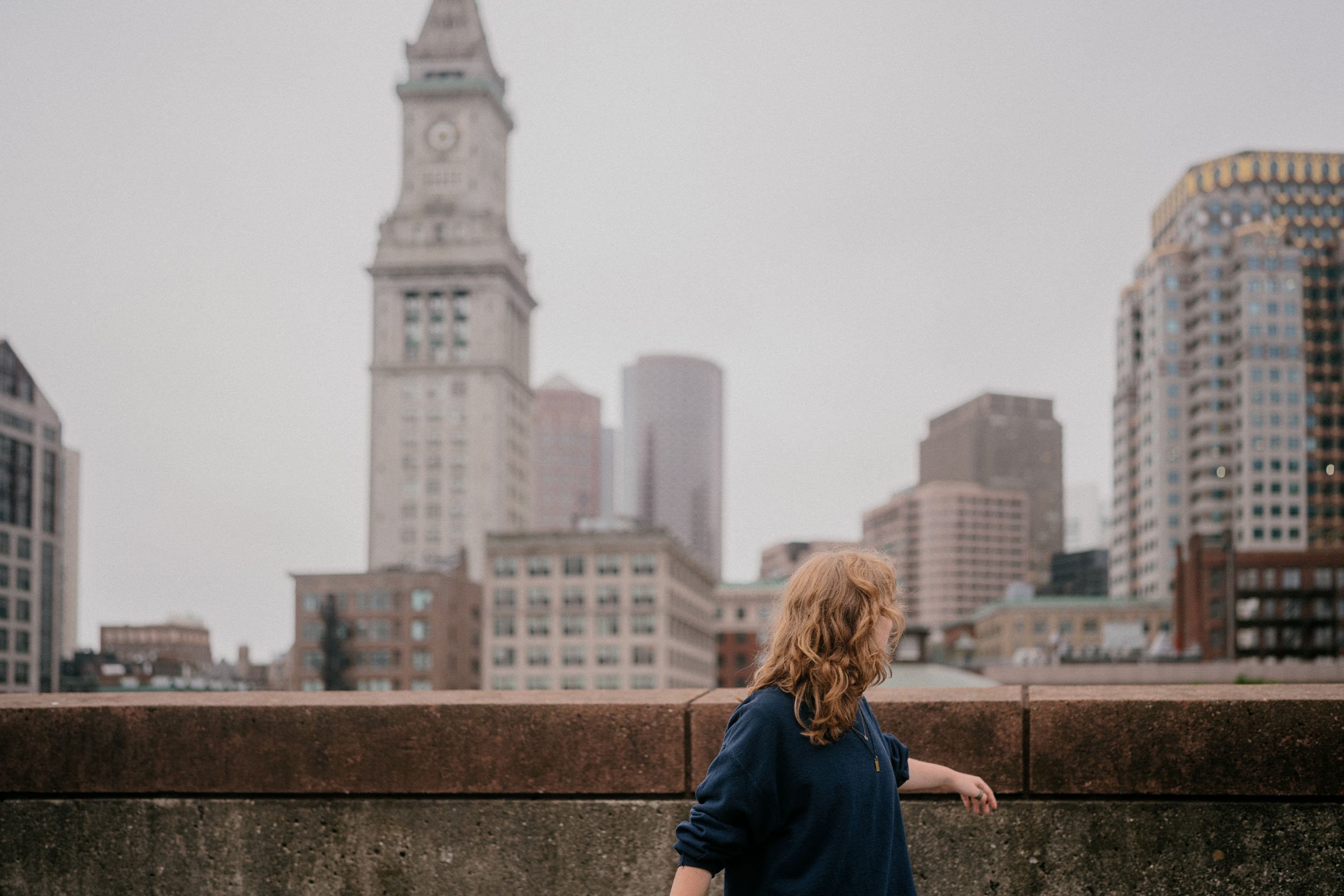 A woman with red hair wearing a navy jacket leaning on a concrete ledge with a city skyline, including a tall clock tower, in the background.