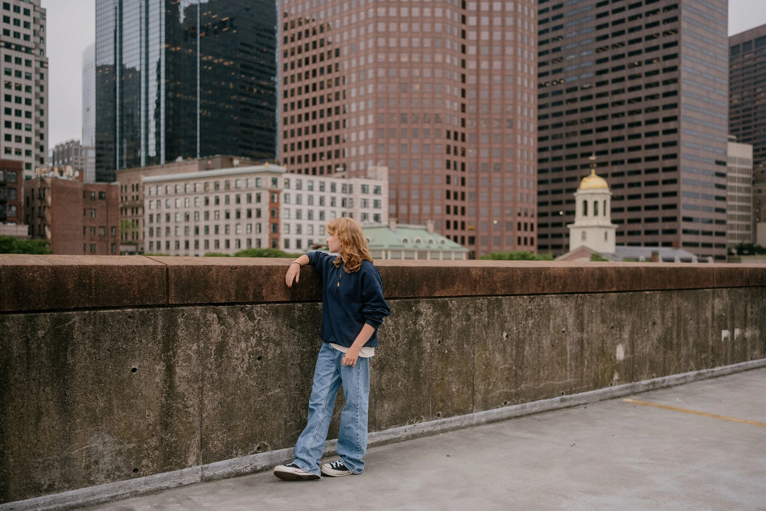 A young person with curly red hair, wearing a navy blue sweatshirt and light blue jeans, leaning against a concrete wall on a rooftop parking lot in an urban cityscape with tall skyscrapers and a church with a gold dome in the background.