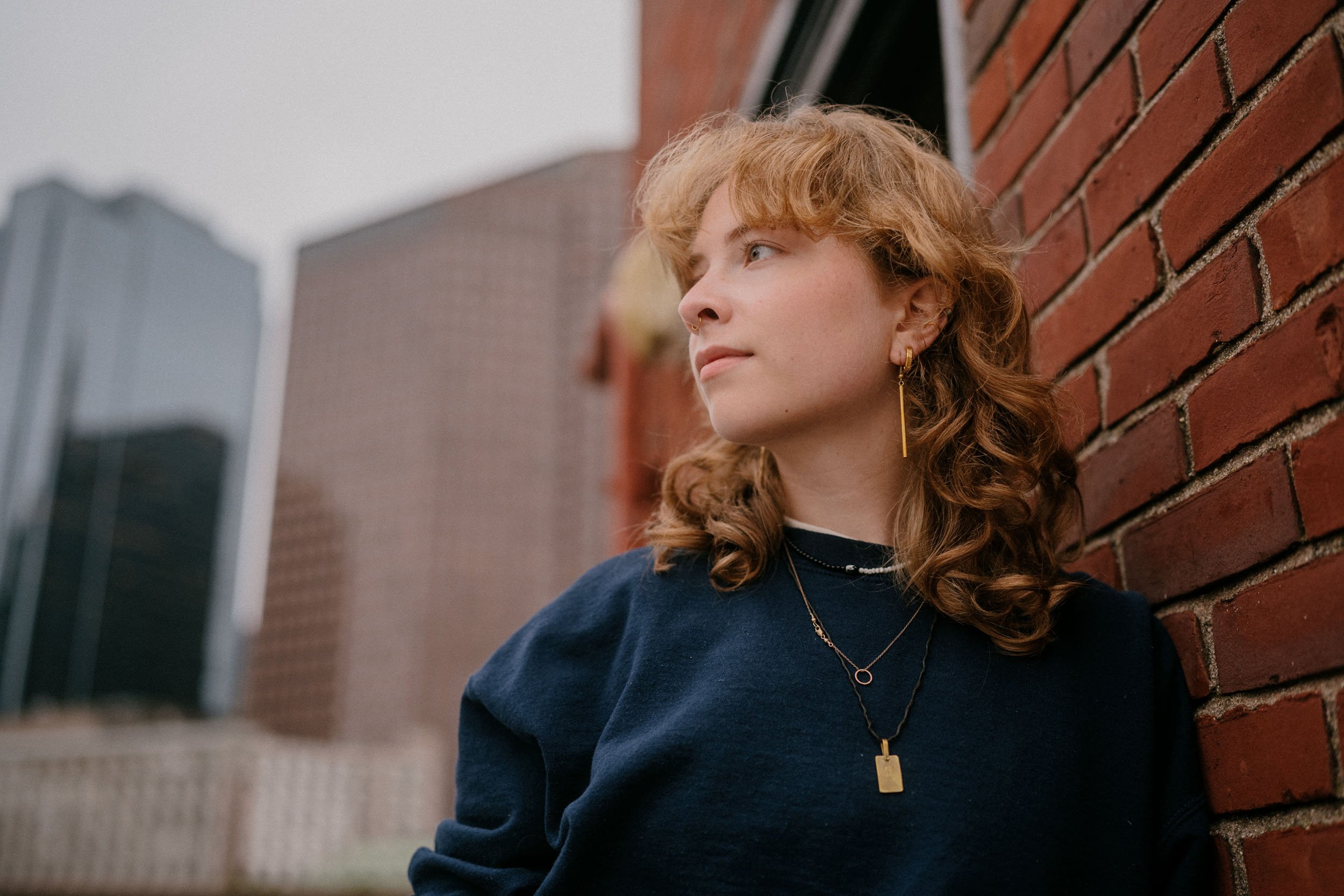 A young woman with curly reddish hair leaning against a brick wall outdoors, looking to the side.