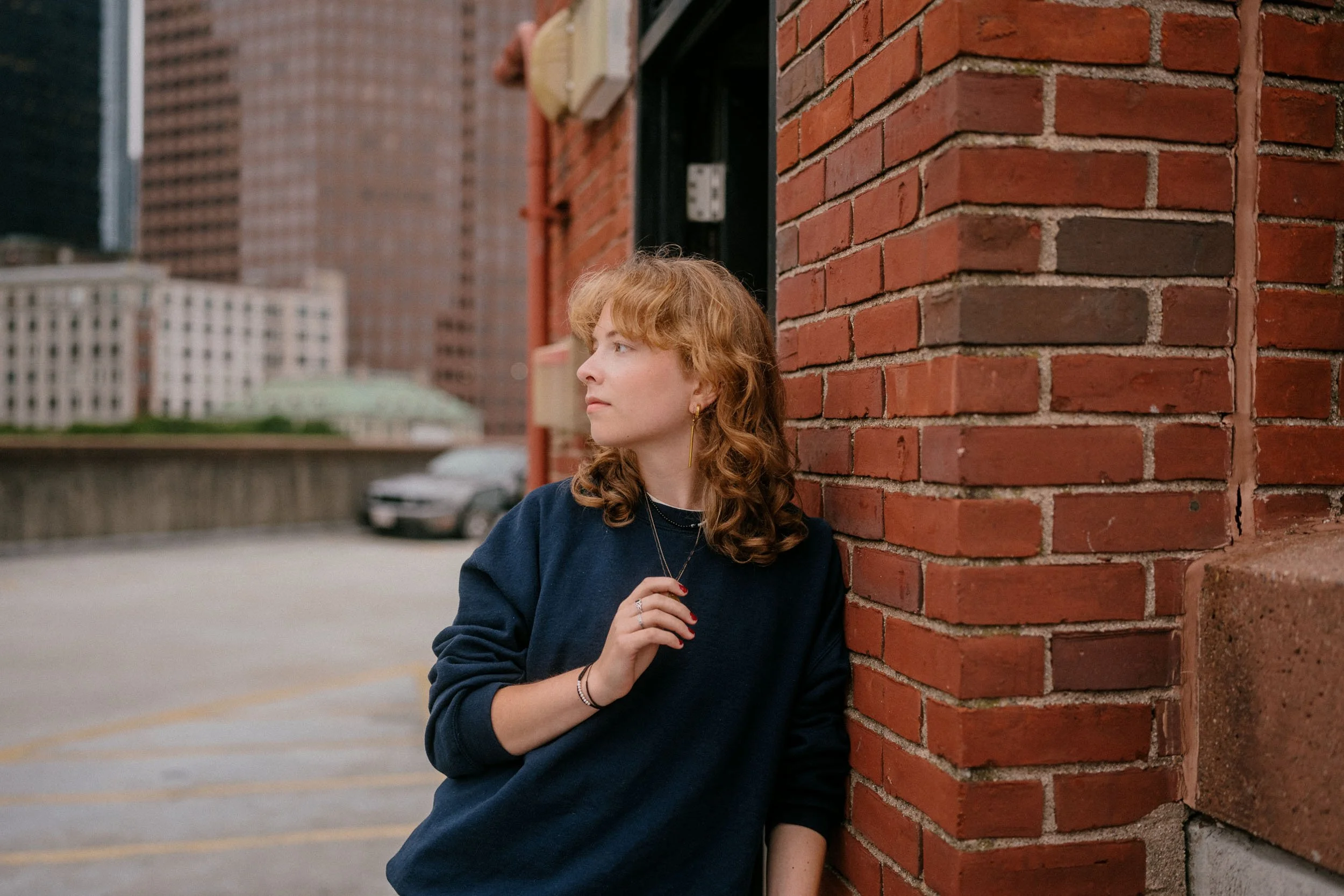 A young woman with curly red hair, wearing a dark blue sweater and jewelry, leaning against a brick wall and looking to her left in an urban setting with tall buildings and a parking lot in the background.