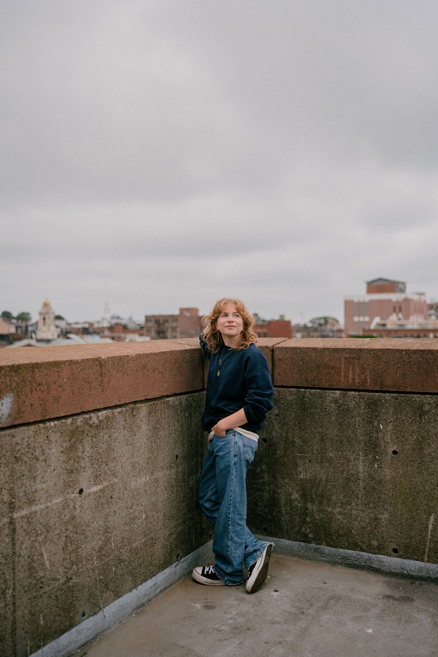 A young woman with curly blonde hair wearing a navy hoodie, jeans, and sneakers standing on a rooftop with city buildings in the background, under an overcast sky.