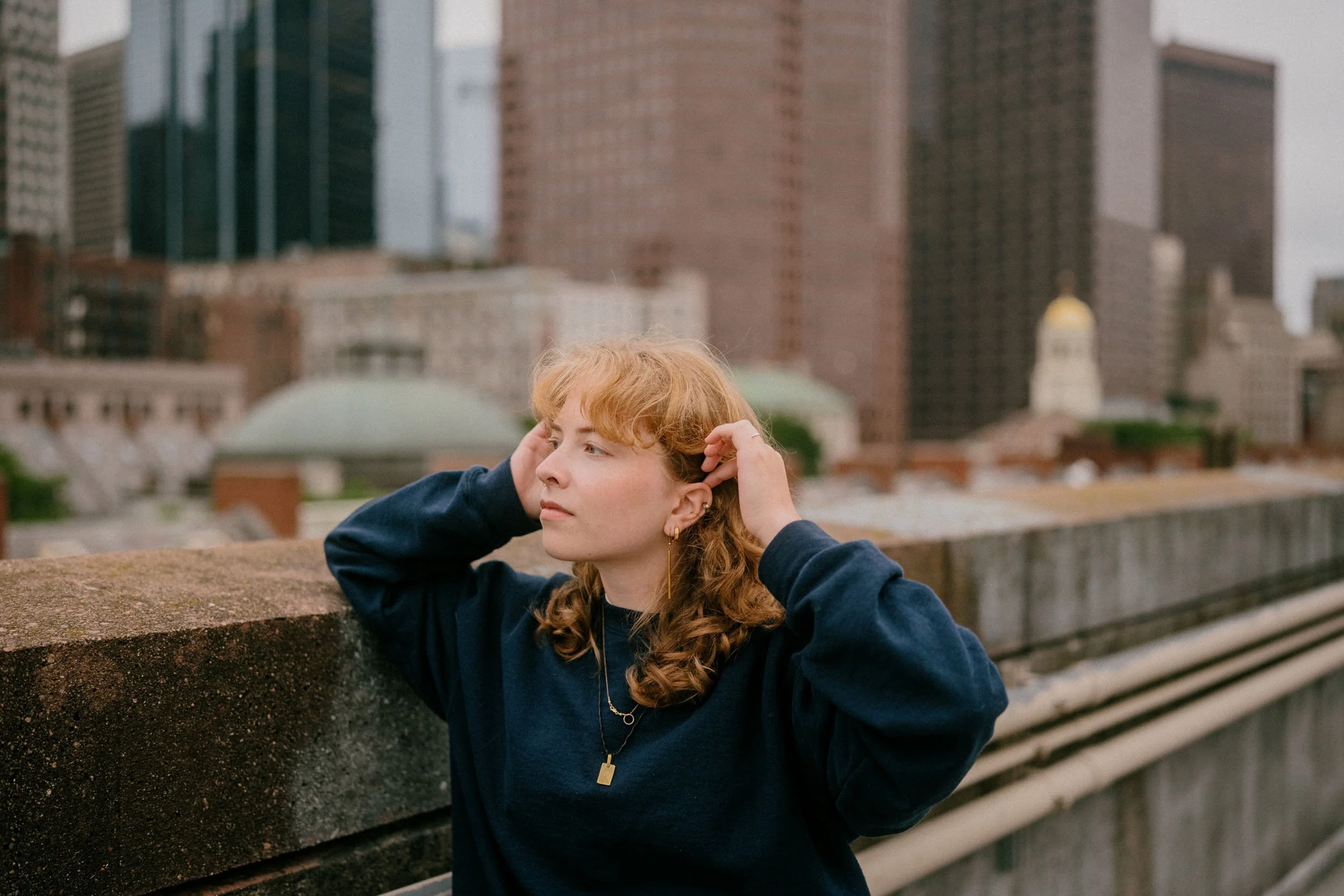 A young woman with reddish curly hair wearing a navy sweatshirt and gold jewelry standing on a city rooftop with buildings and a cloudy sky in the background.