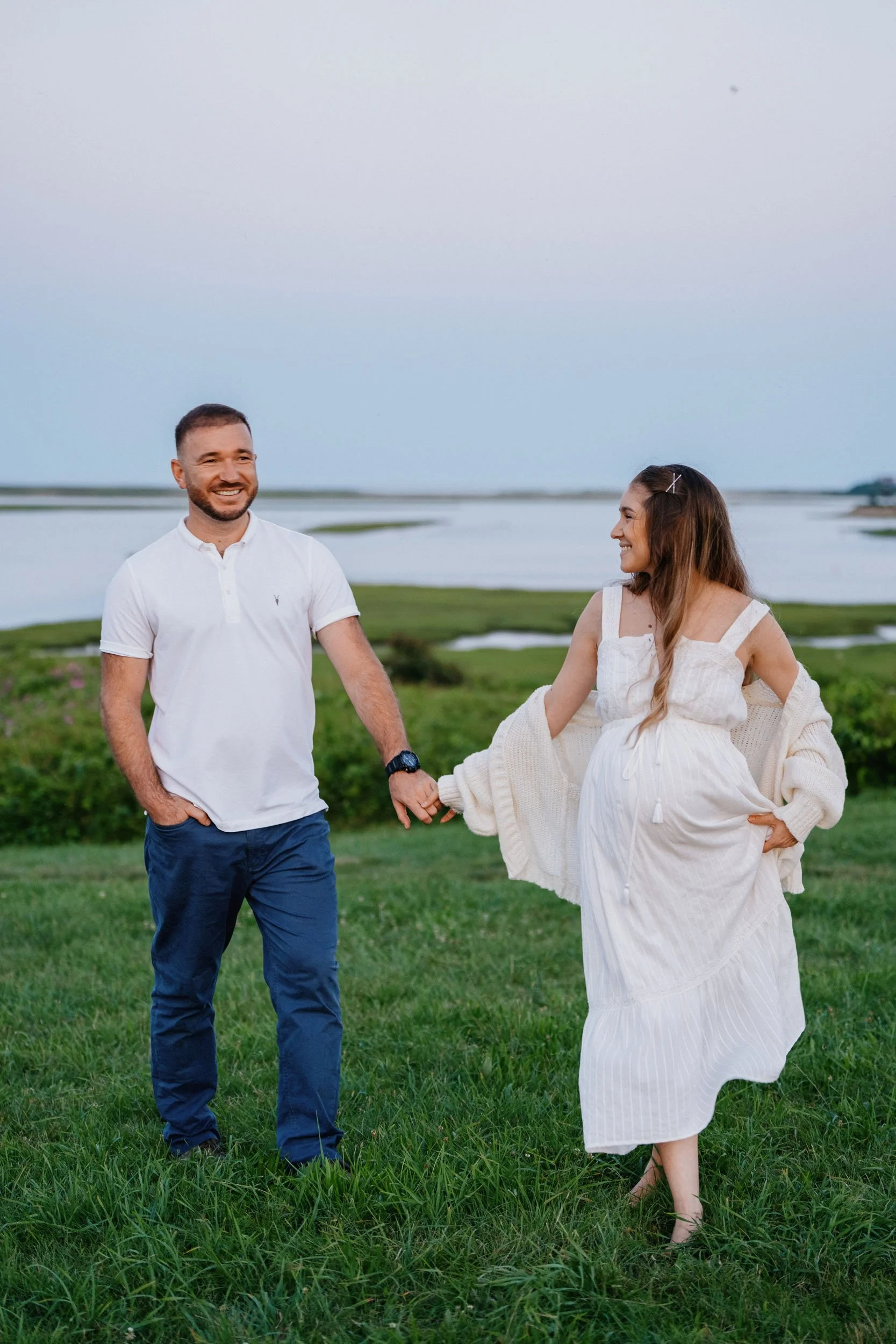 A smiling couple holding hands and walking on grassy land near a body of water with a distant shoreline, during what appears to be late afternoon or early evening.