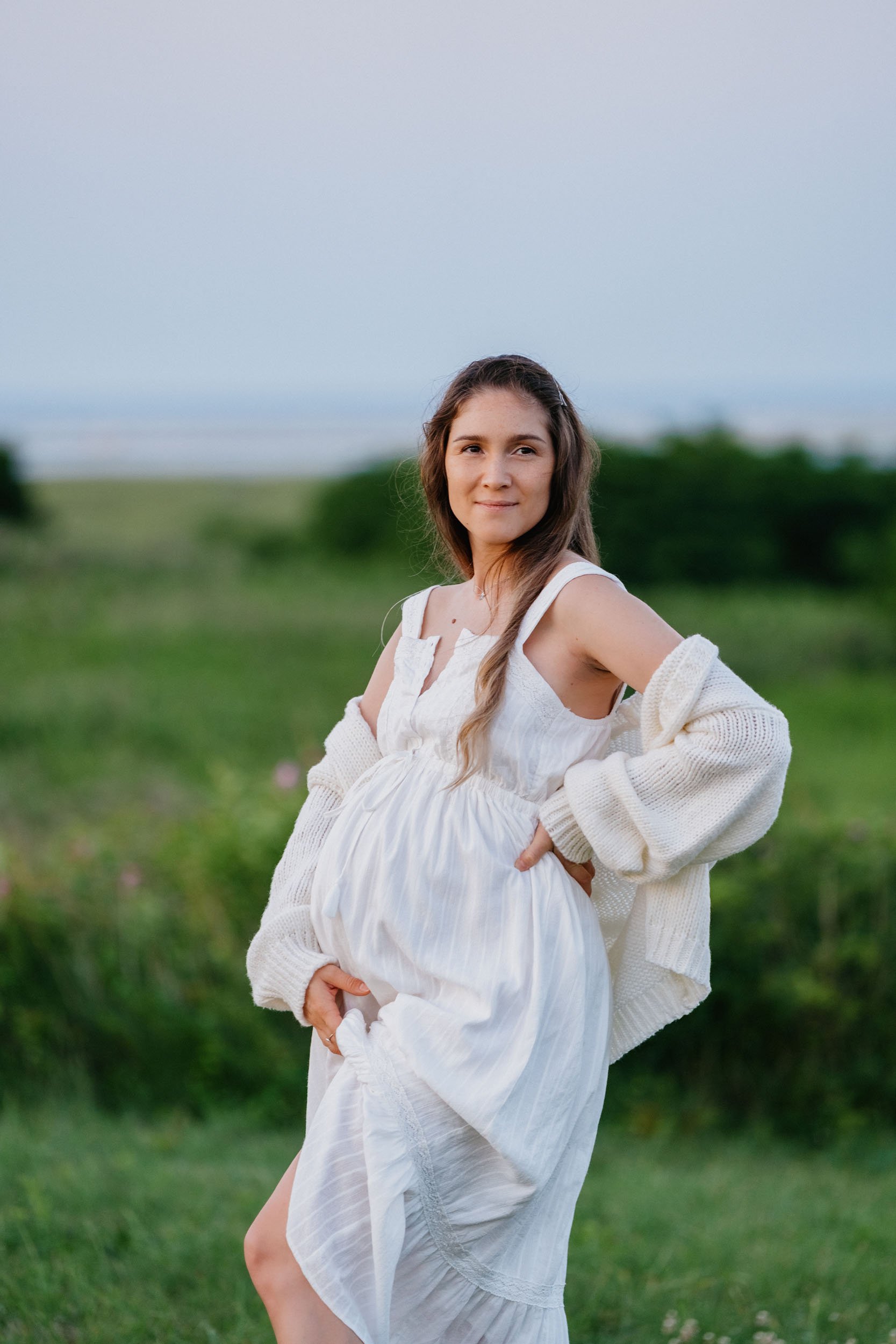 A pregnant woman in a white dress and cardigan standing outdoors on a grassy field with trees in the background.