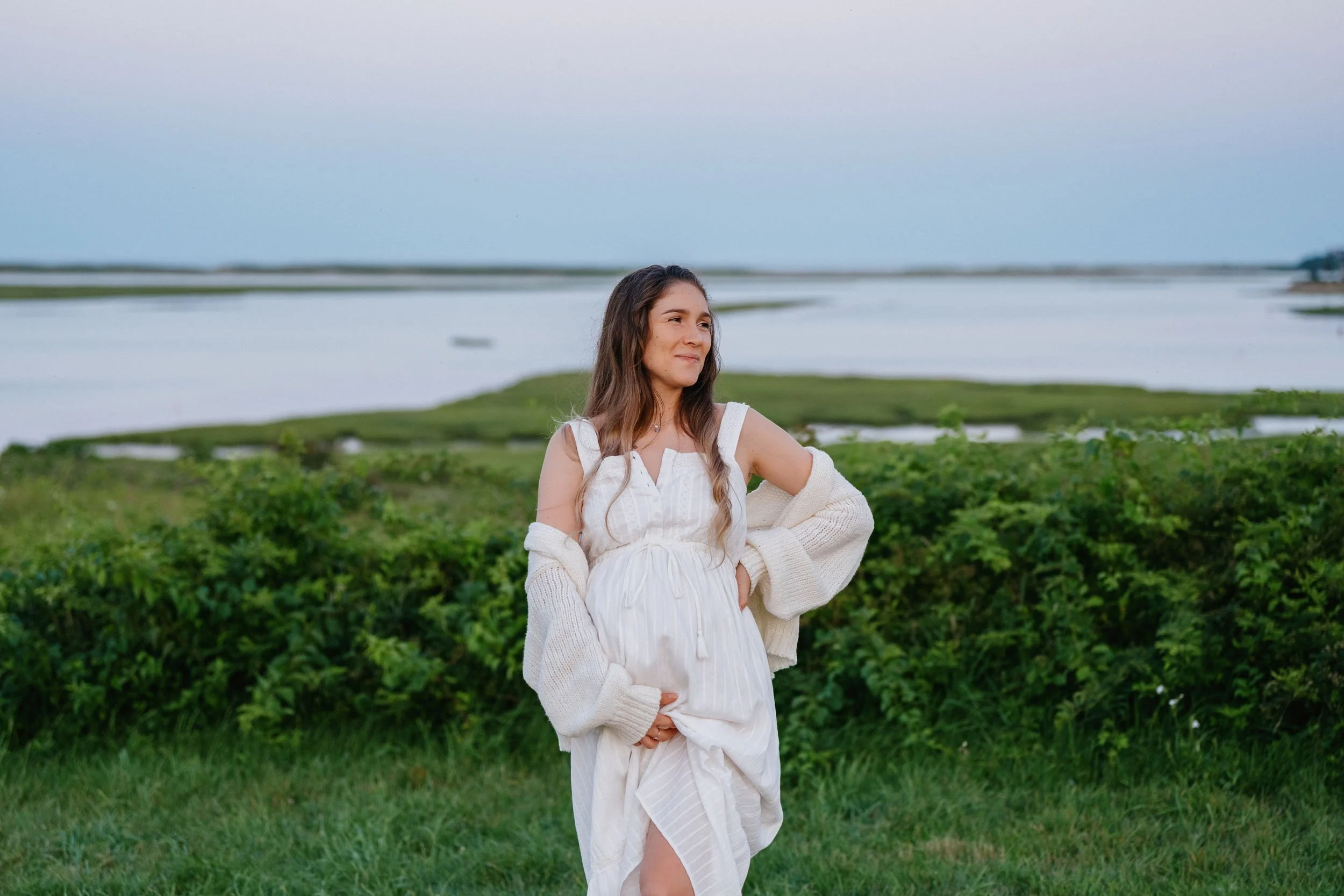 A young woman in a white dress and cream cardigan standing in a grassy area near a lake or river with green bushes and a cloudy sky in the background.