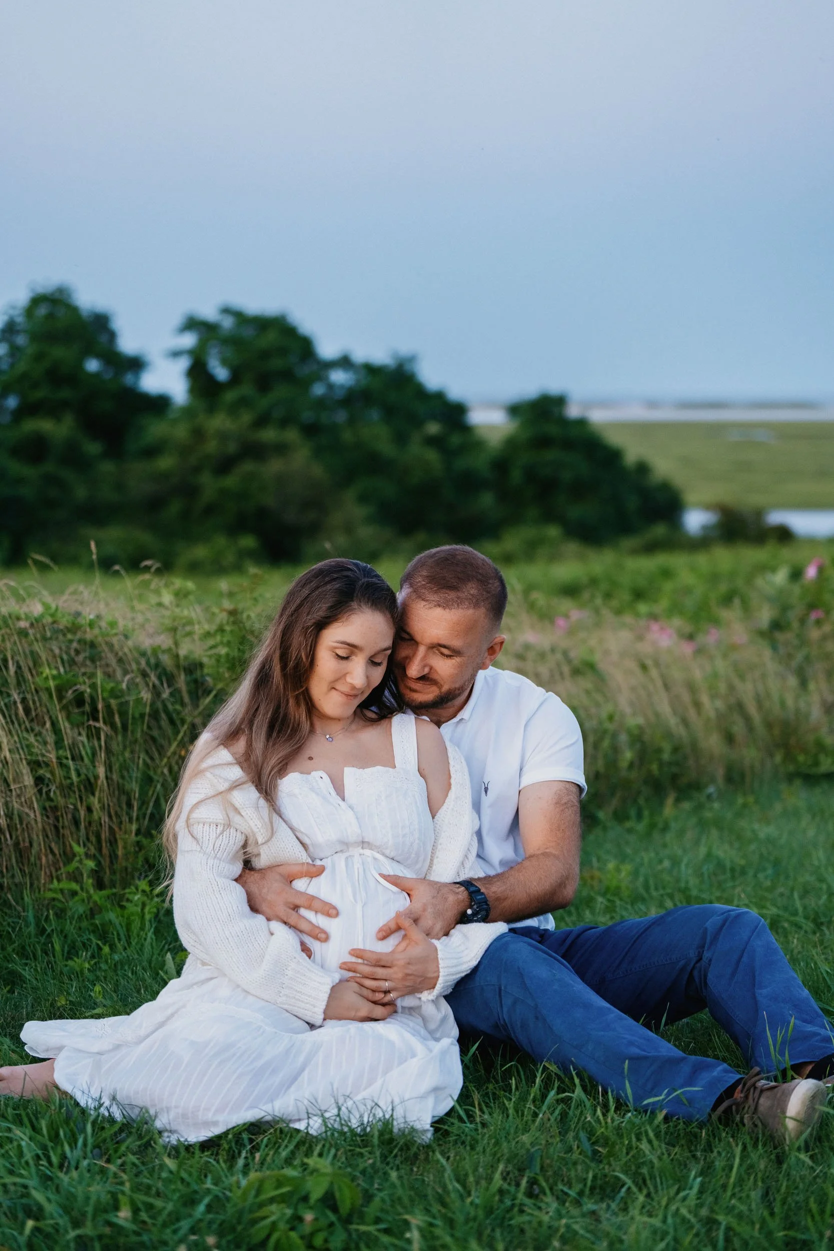 A pregnant woman and her partner sitting on grass in a field, embracing and smiling with trees and a body of water in the background during dusk.