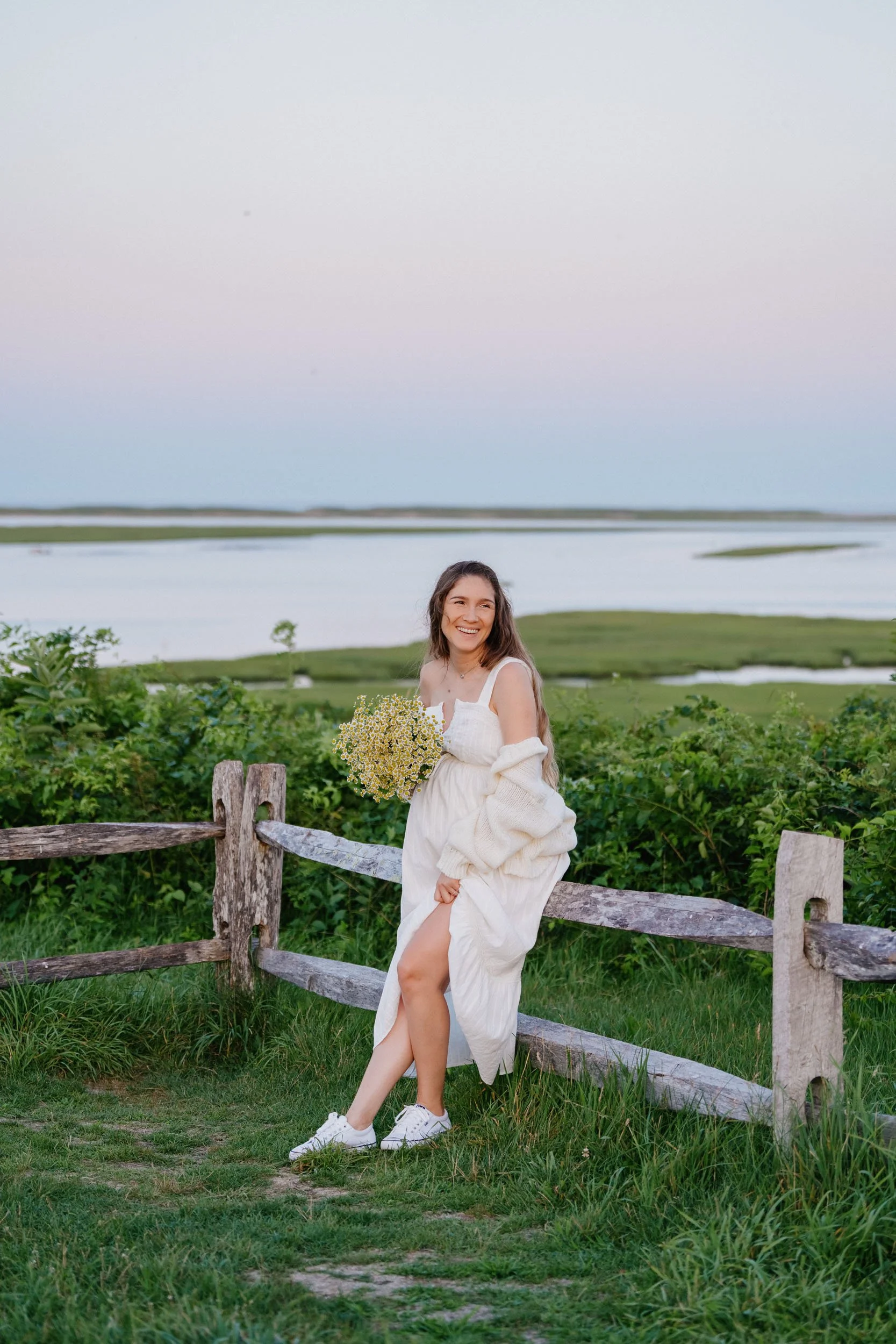 A young woman in a white dress and sneakers holding a bouquet of yellow flowers, sitting on a wooden fence in a grassy field near a body of water with wetlands, under a pastel-colored sky.