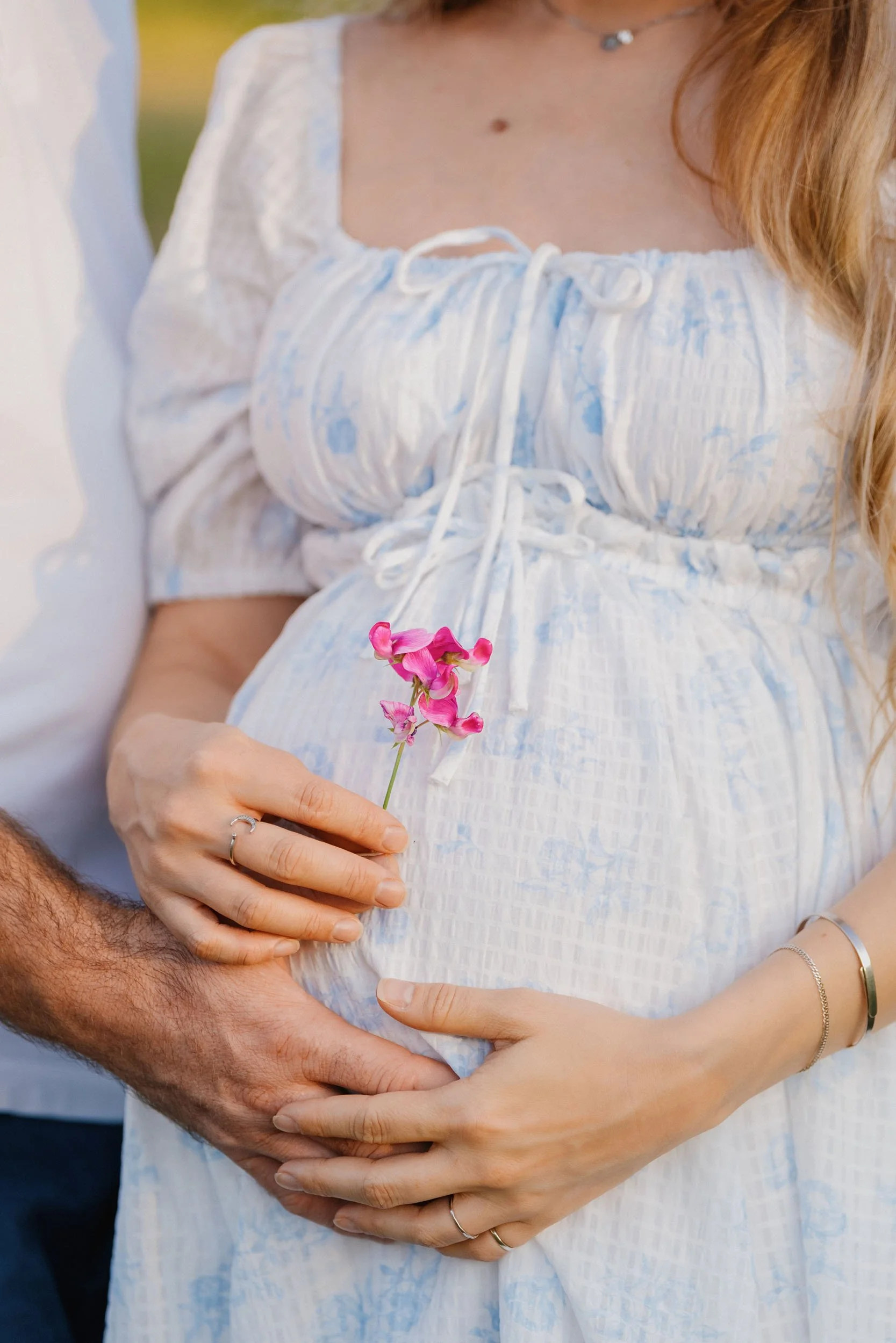 Close-up of a pregnant woman holding a pink flower with a man's hand gently placed on her belly.