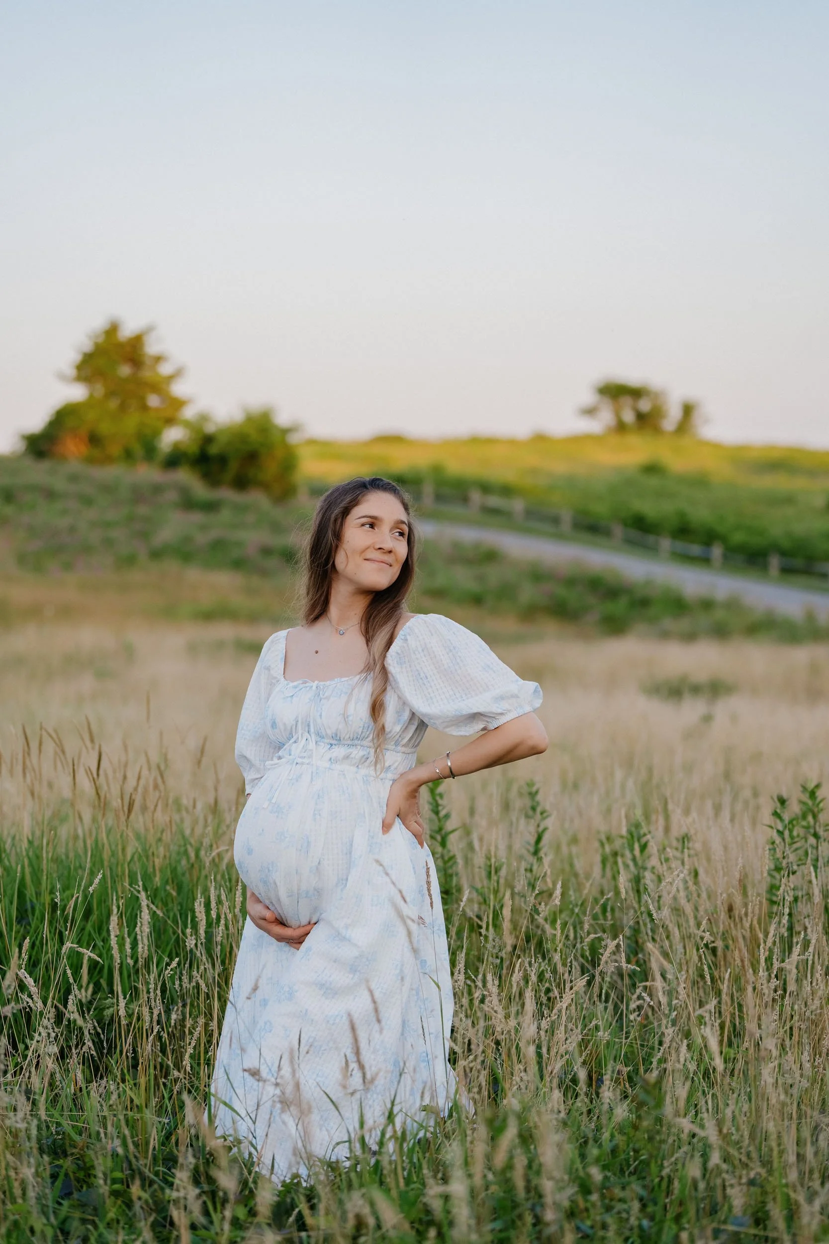 Pregnant woman standing in a grassy field with her hand on her belly, wearing a white dress, during sunset.