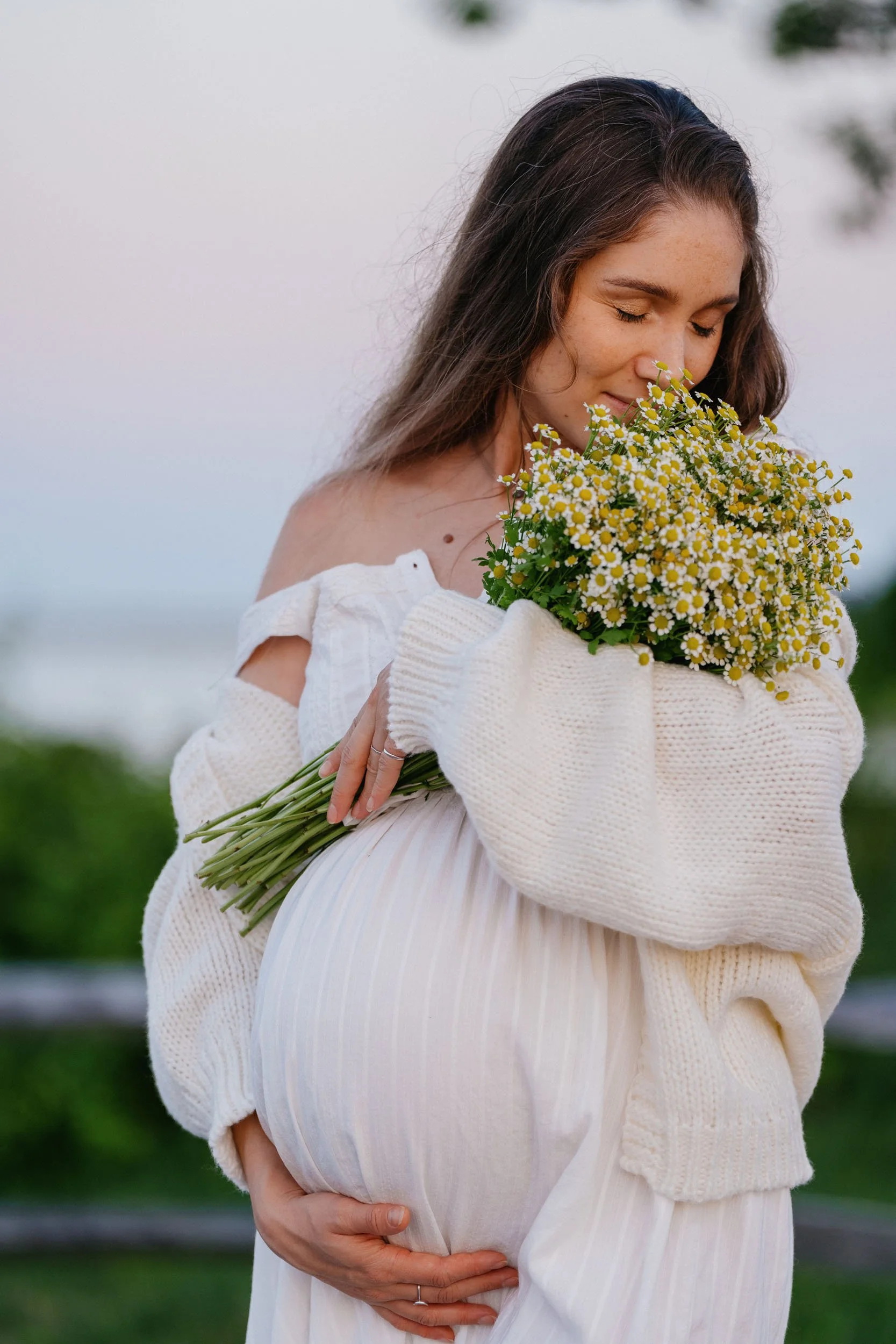 A pregnant woman with long brown hair, holding a bouquet of white and yellow daisies, standing outdoors during sunset, wearing a white off-shoulder dress and a cozy cream sweater.