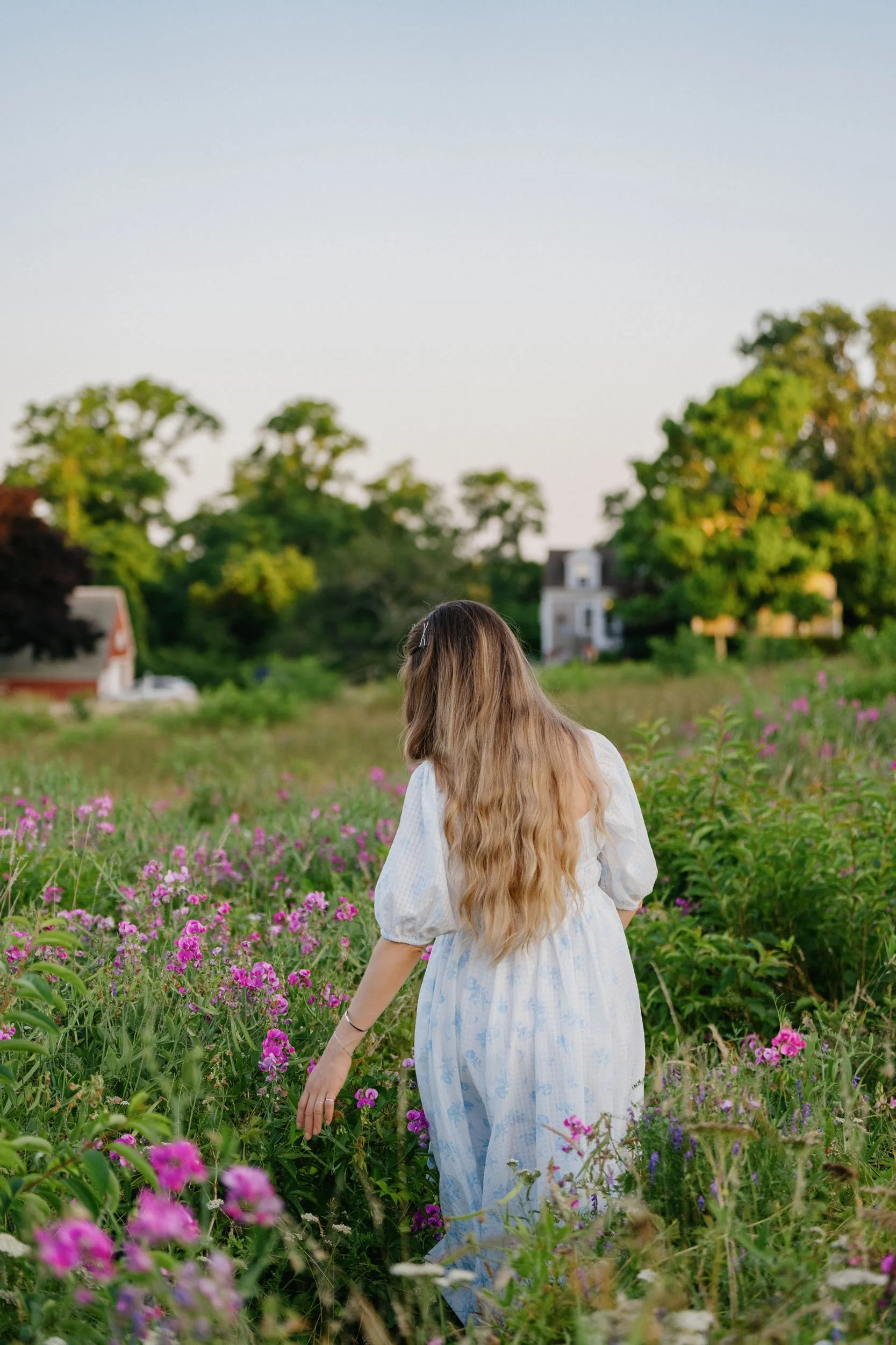 A woman with long, wavy blonde hair, wearing a white dress with puffy sleeves, walking through a field of pink and purple flowers on a clear day with a few houses and green trees in the background.