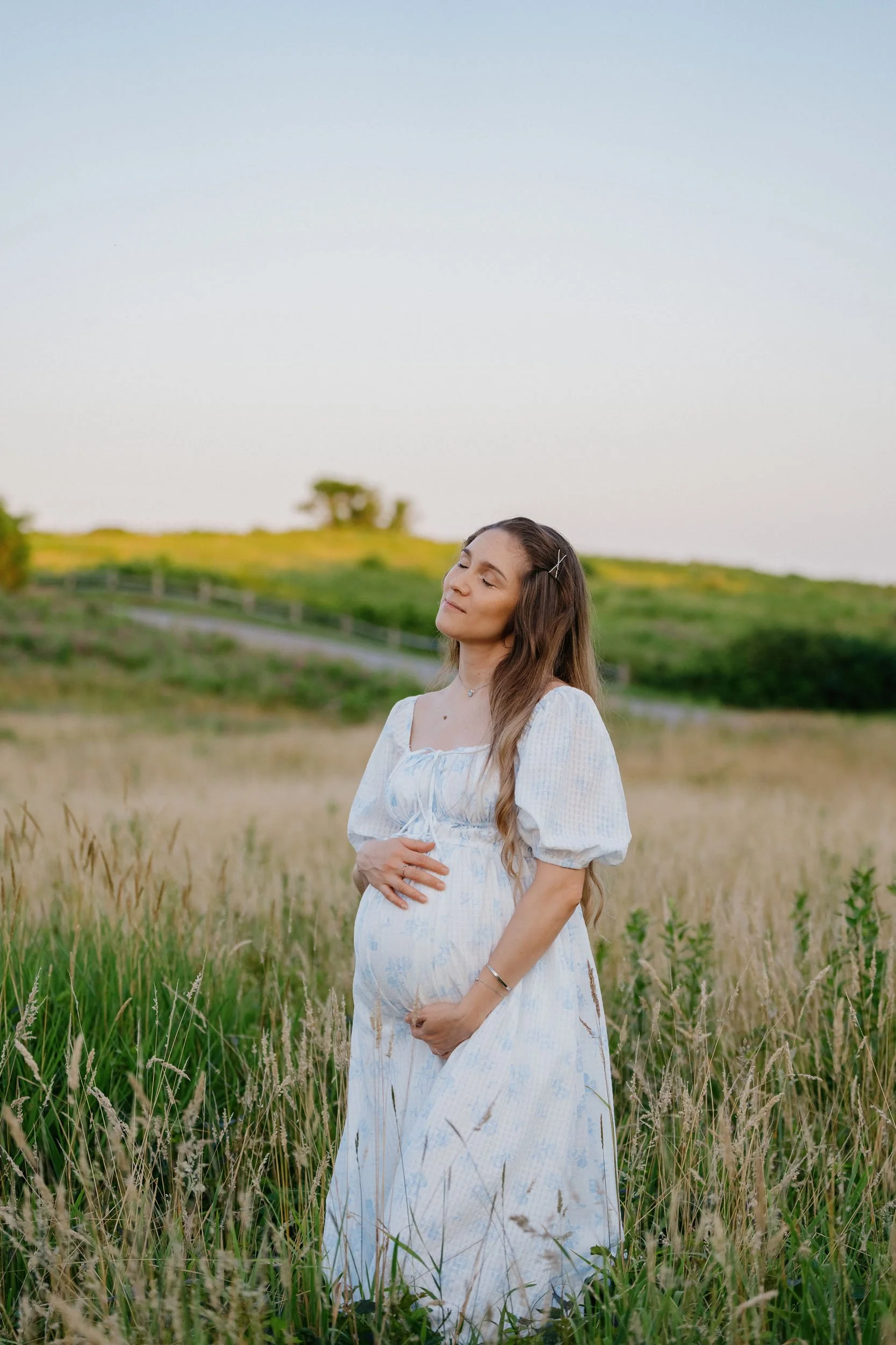 A pregnant woman in a white dress standing in a field of tall grass with her eyes closed, holding her belly, with green rolling hills and a clear sky in the background.