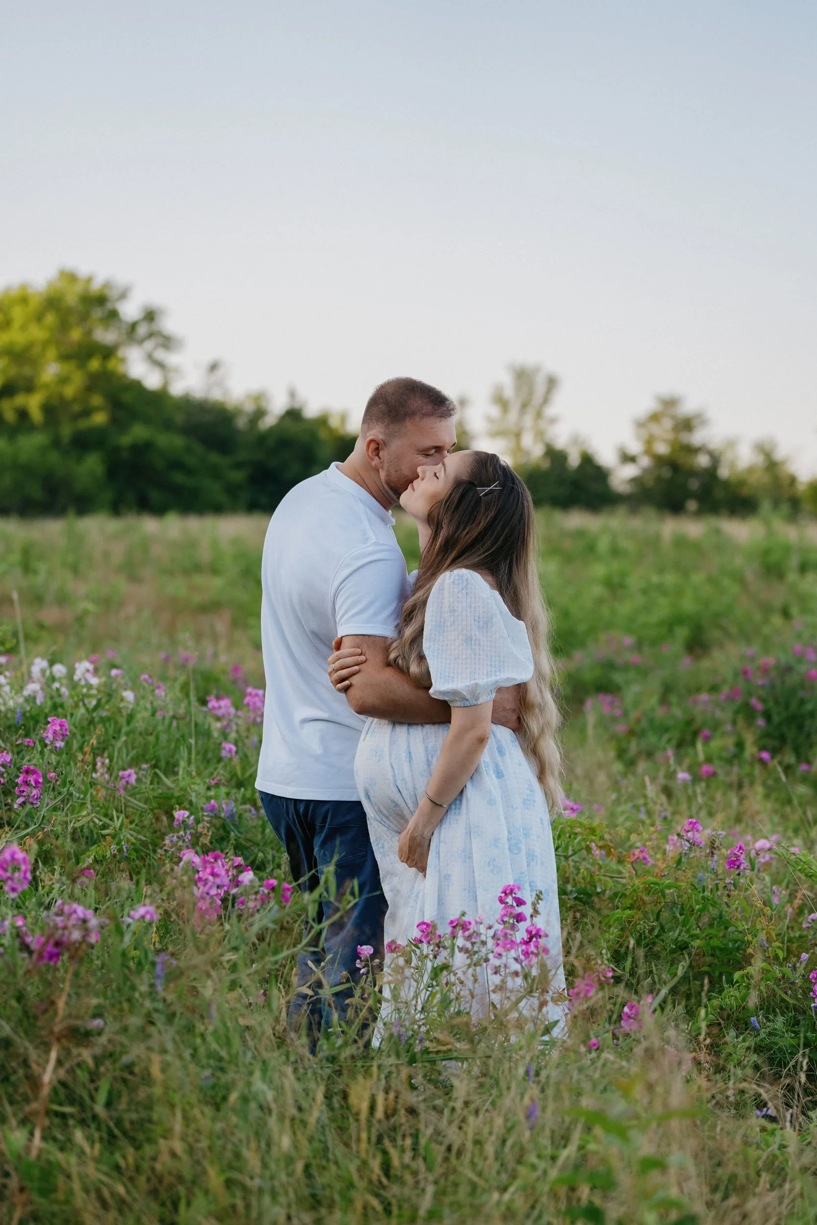 A couple sharing a kiss in a field of pink and purple flowers with trees in the background.