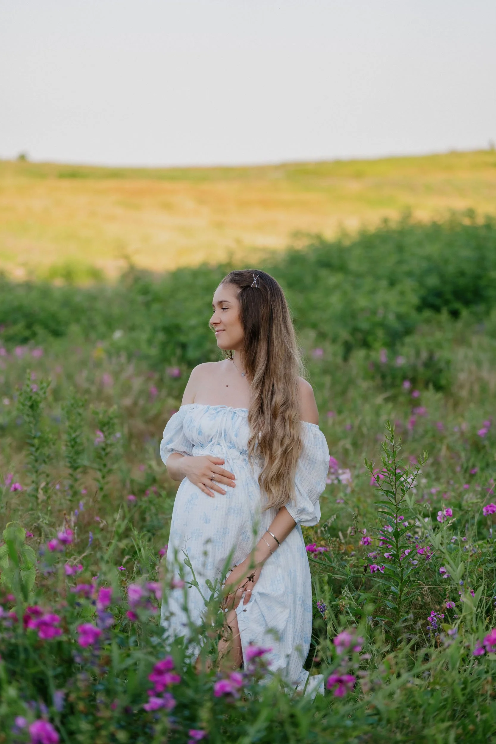 A pregnant woman with long wavy hair wearing a white off-shoulder dress, standing in a field of pink and purple flowers with green shrubs and a yellow hillside in the background.