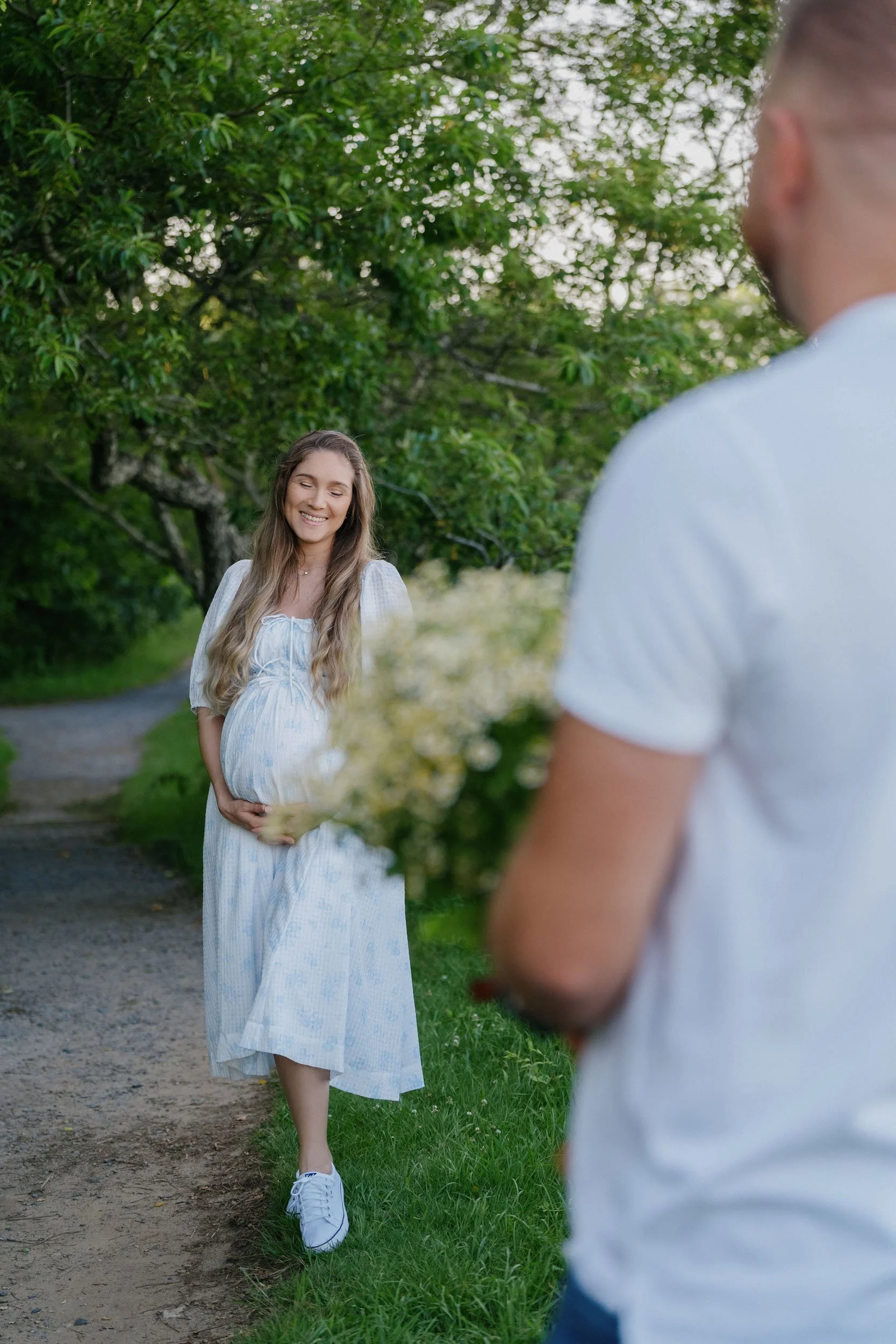 A pregnant woman in a white dress holding flowers and smiling at a man, who is holding flowers, outdoors with green trees in the background.