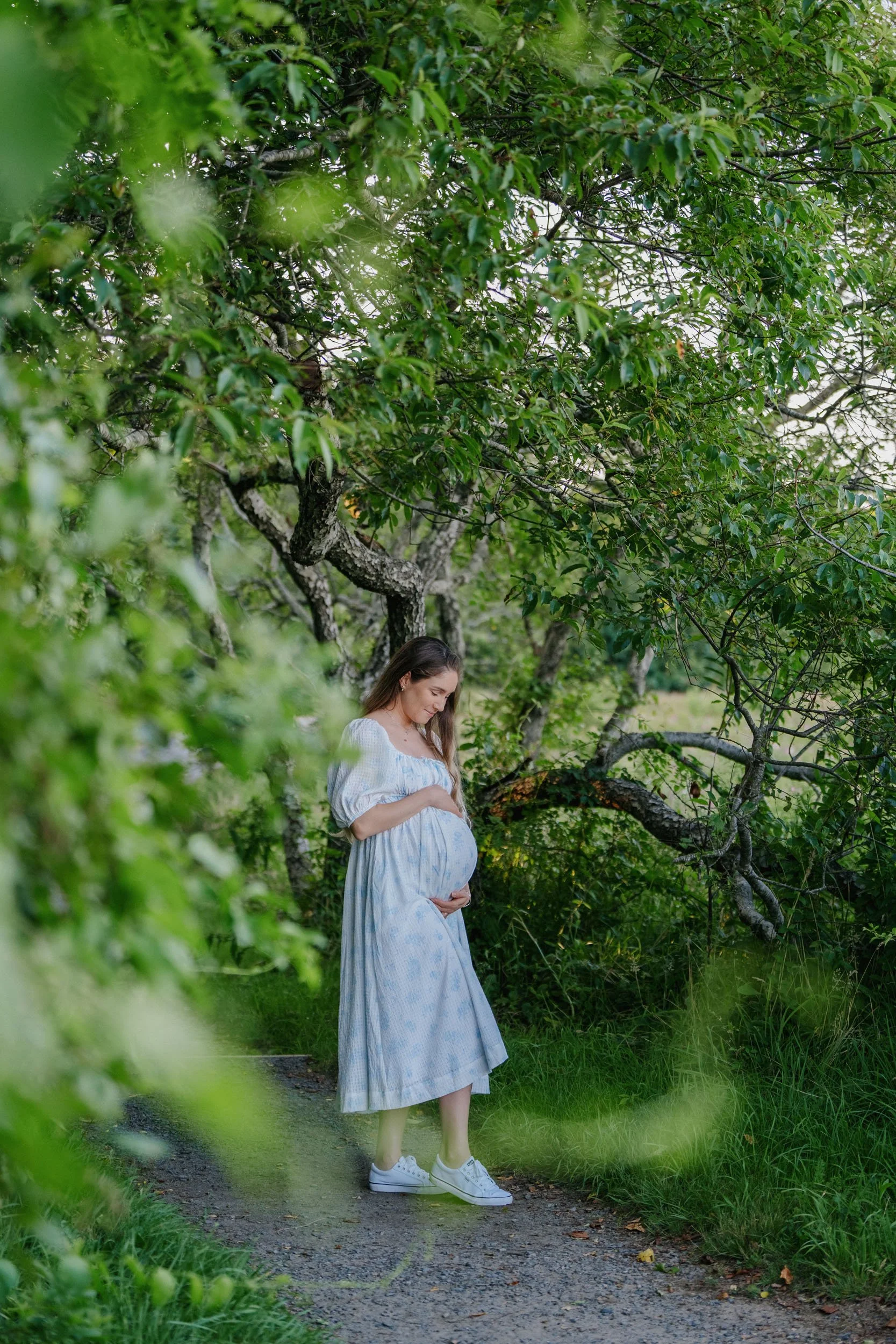 Pregnant woman standing on a wooded path, looking down and holding her belly, surrounded by lush green foliage.