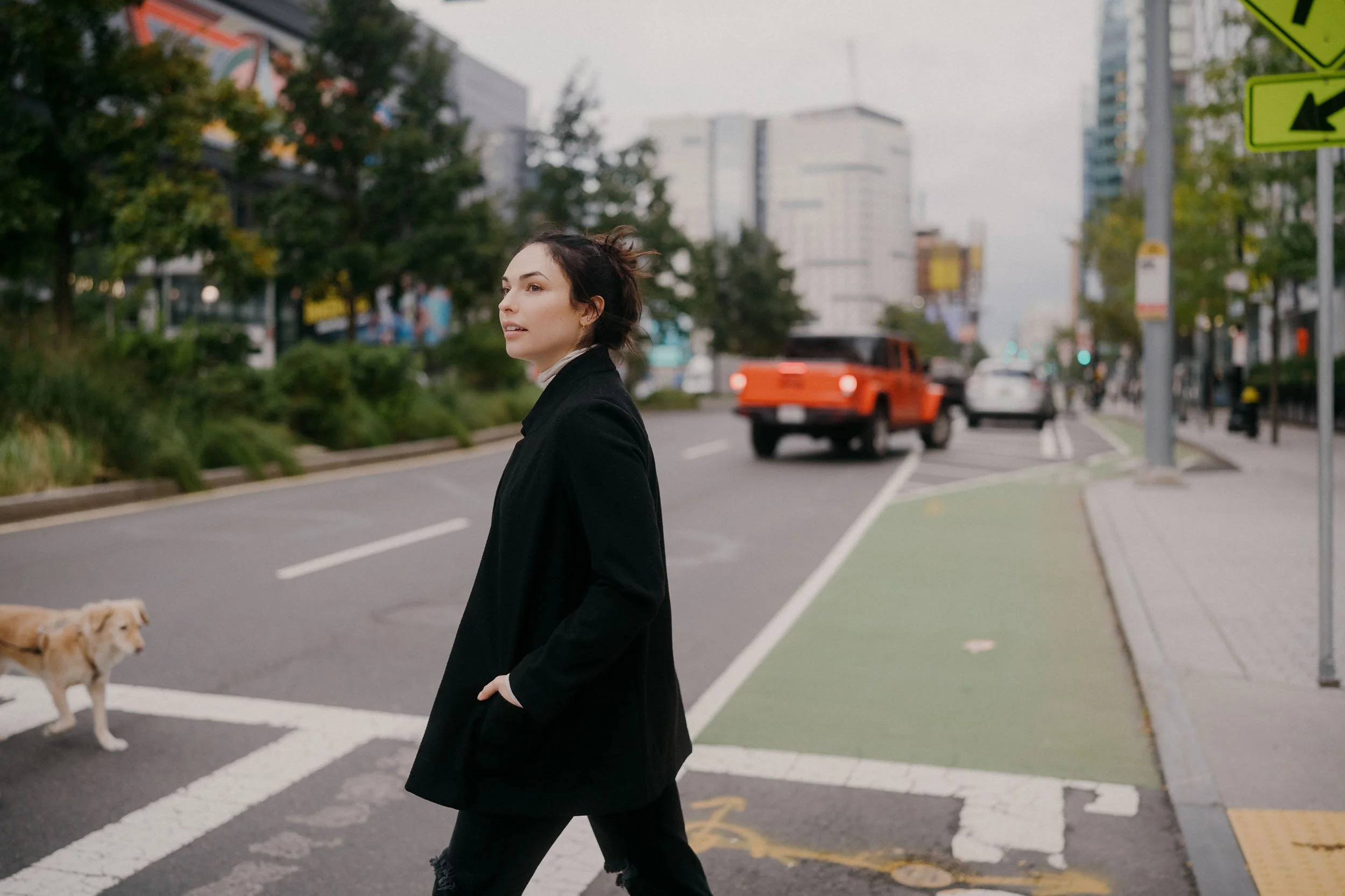 A woman with dark hair in a black coat walking across a crosswalk in an urban area with buildings, cars, and a dog nearby.