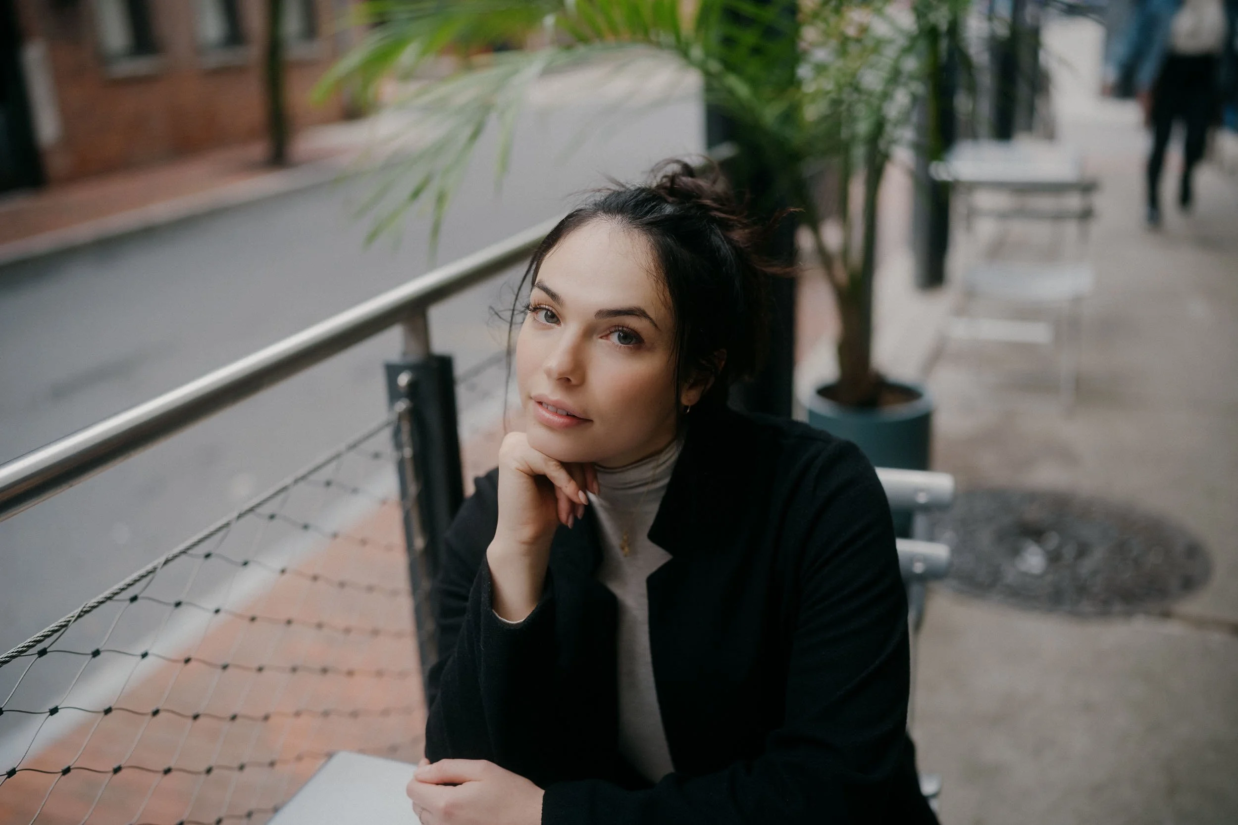 A young woman sitting at an outdoor cafe or patio, resting her chin on her hand, looking at the camera with a slight smile, with plants and an urban street in the background.