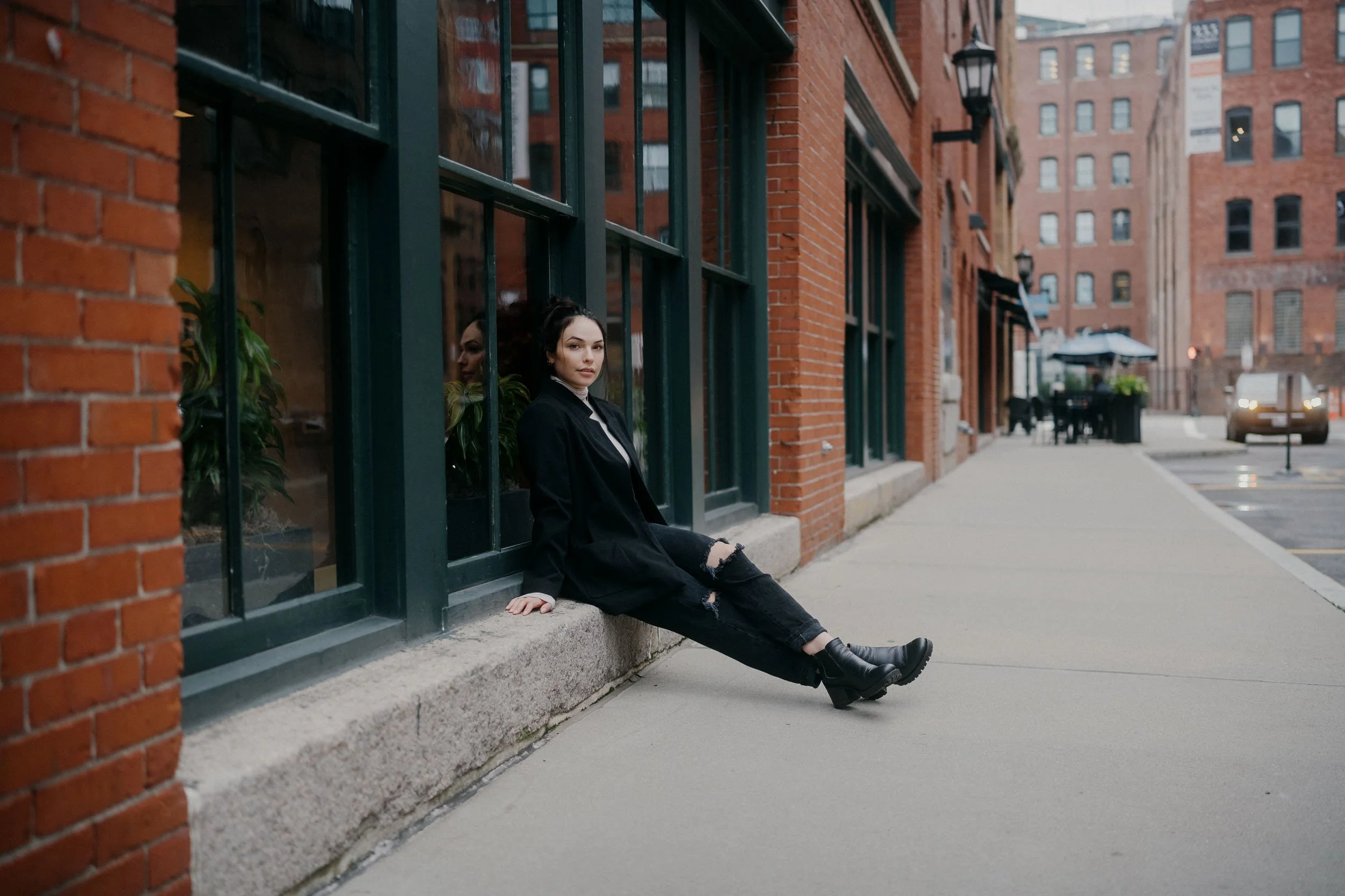 A young woman with dark curly hair, wearing a black jacket, ripped black jeans, and black boots, sits on a sidewalk outside a red brick building with large green window frames. She is leaning back against the window ledge, looking at the camera with 