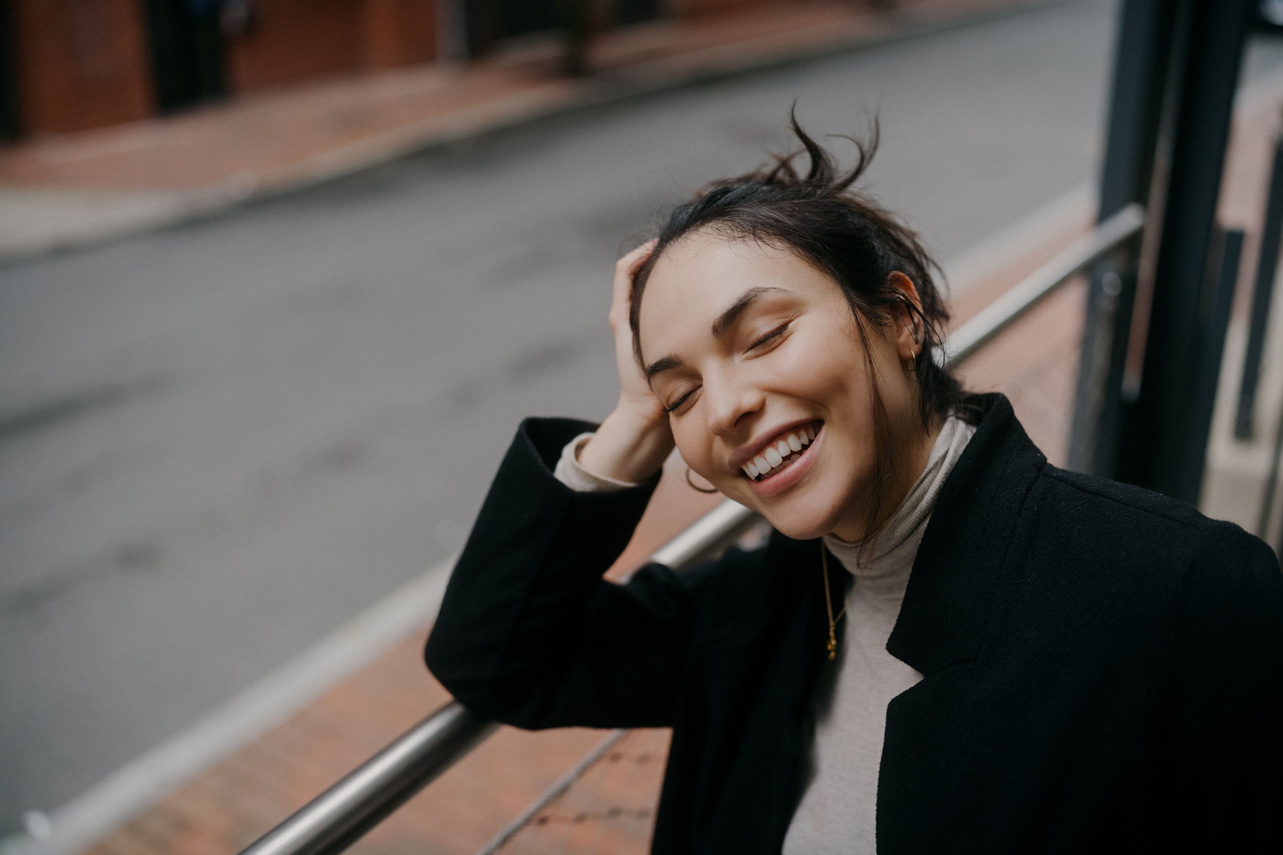 A woman smiling happily with her eyes closed, resting her head on her hand, outdoors on a city sidewalk.