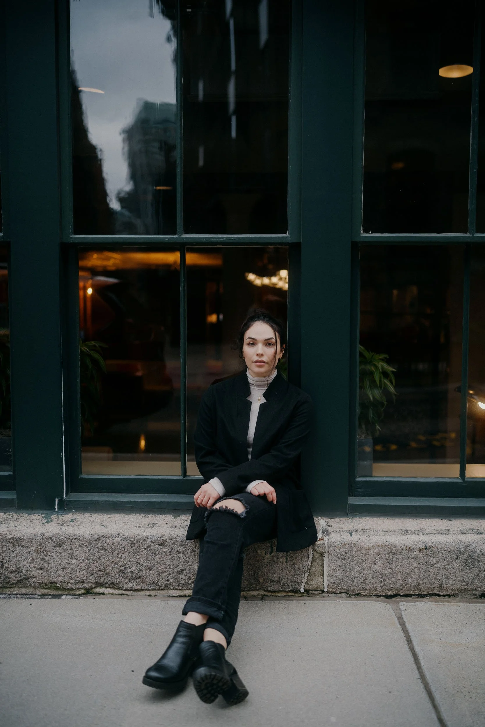 A young woman sitting on a concrete sidewalk outside a building with large, dark green framed windows at dusk or evening.