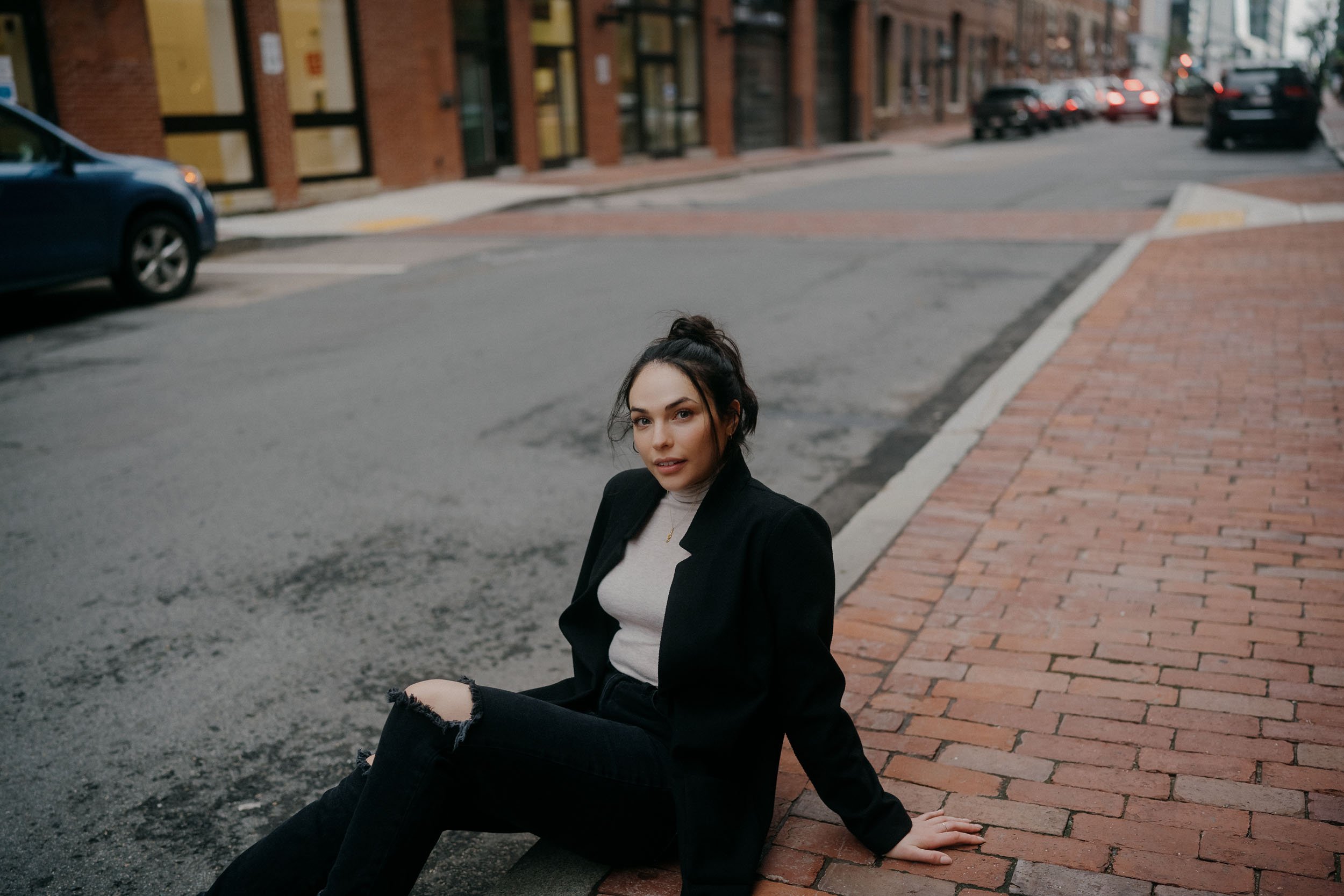 A young woman with dark hair tied up in a bun, wearing a black blazer and ripped black jeans, sitting on a brick sidewalk next to a street with cars and buildings in the background.