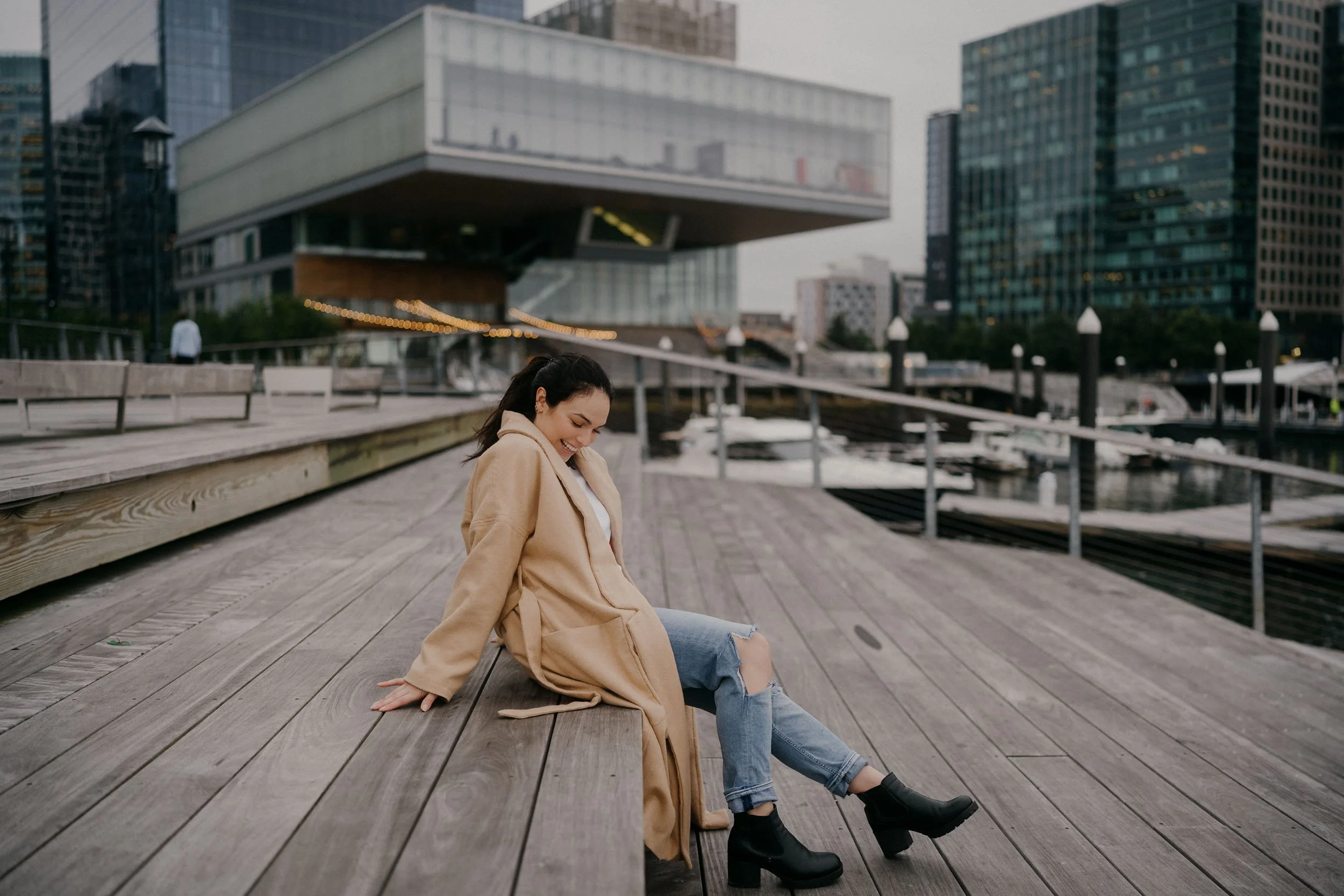 A woman with long dark hair, wearing a beige coat, ripped jeans, and black ankle boots, sitting on a wooden dock near a marina with boats, city buildings, and a modern architectural structure in the background. The woman is smiling and looking down.